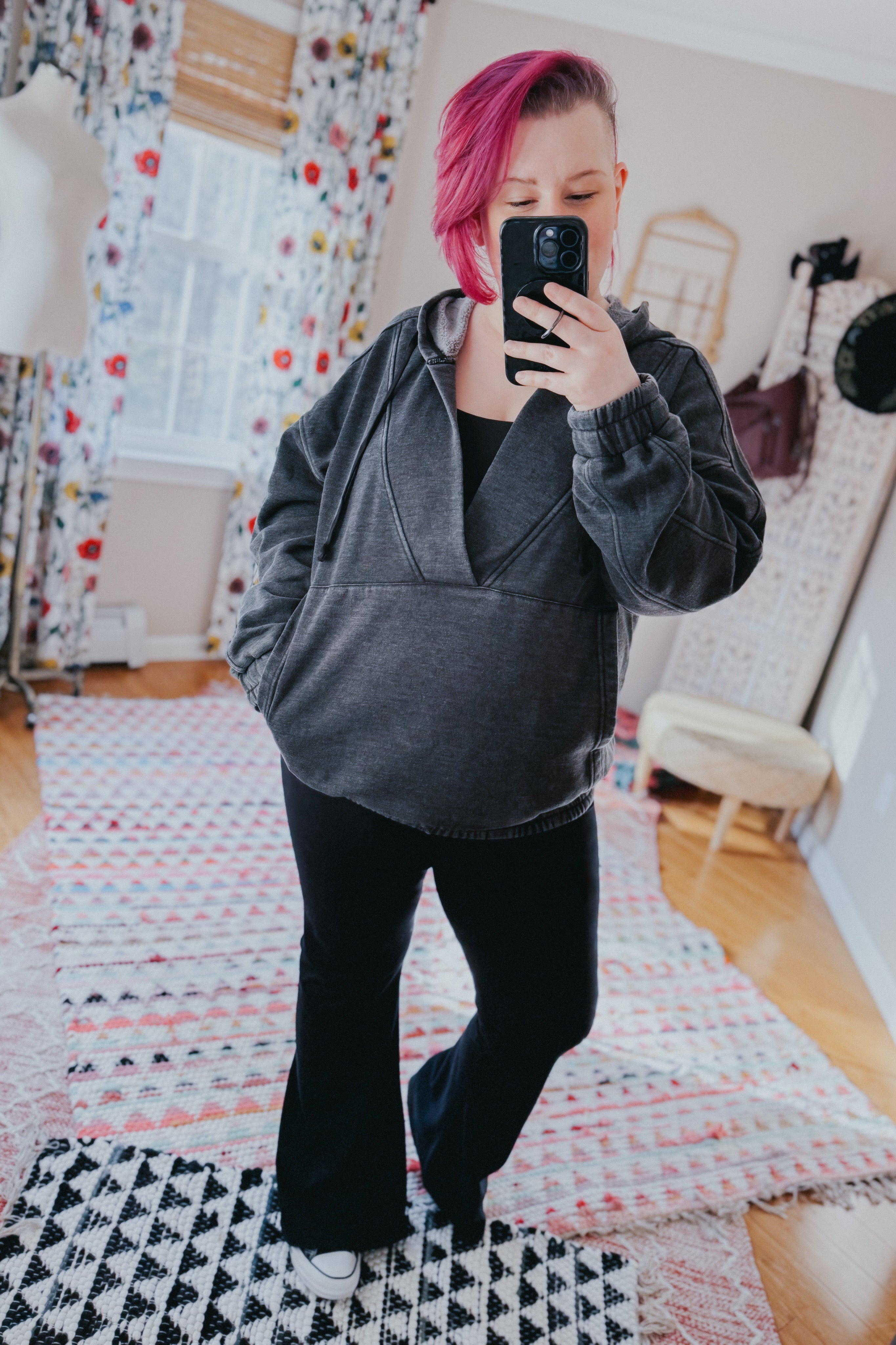Woman with pink hair wearing charcoal acid wash hoodie and black pants taking indoor mirror selfie in bright room