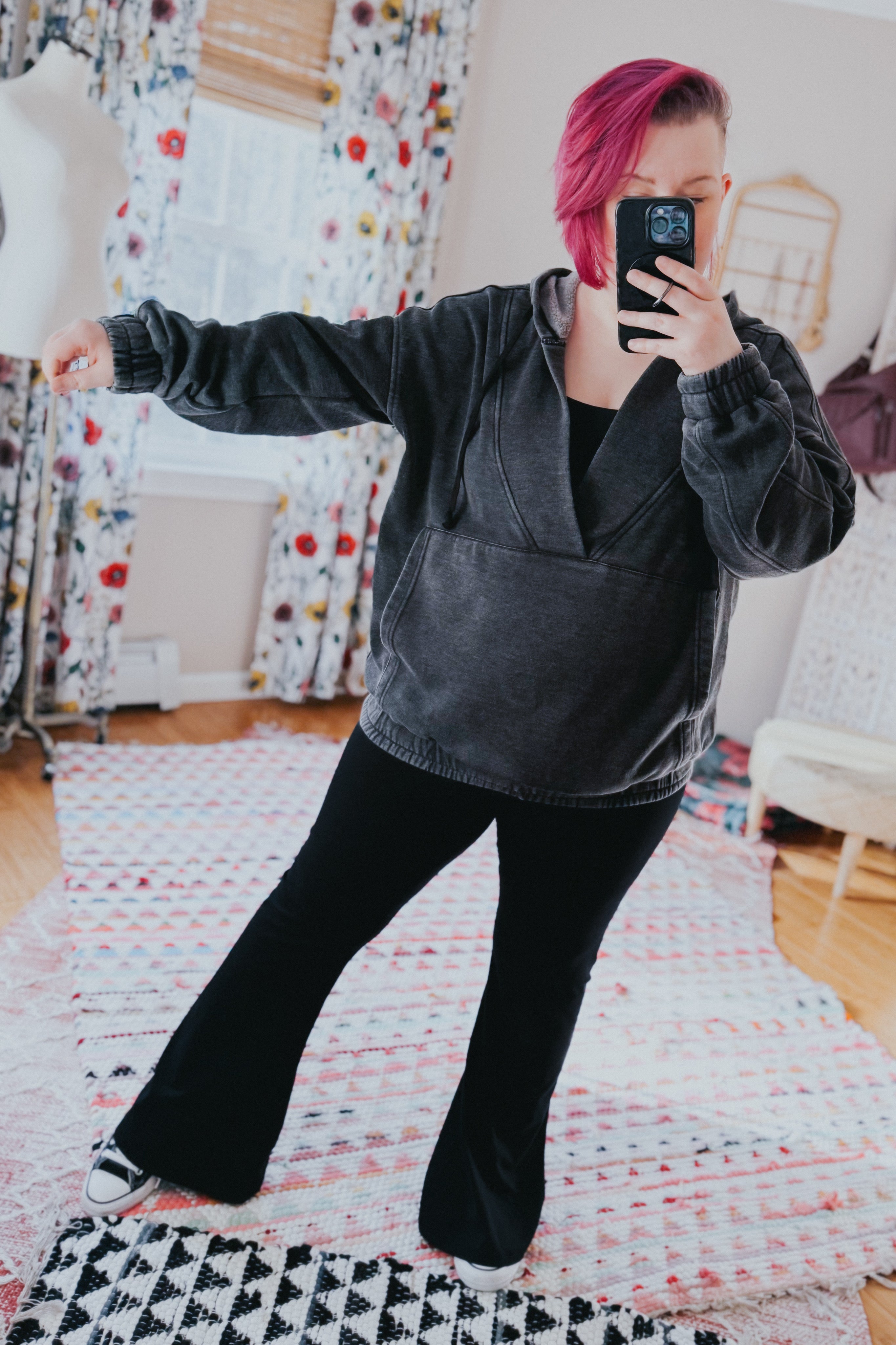 Woman with short magenta hair wearing charcoal acid wash hoodie and black pants taking mirror selfie in bright room