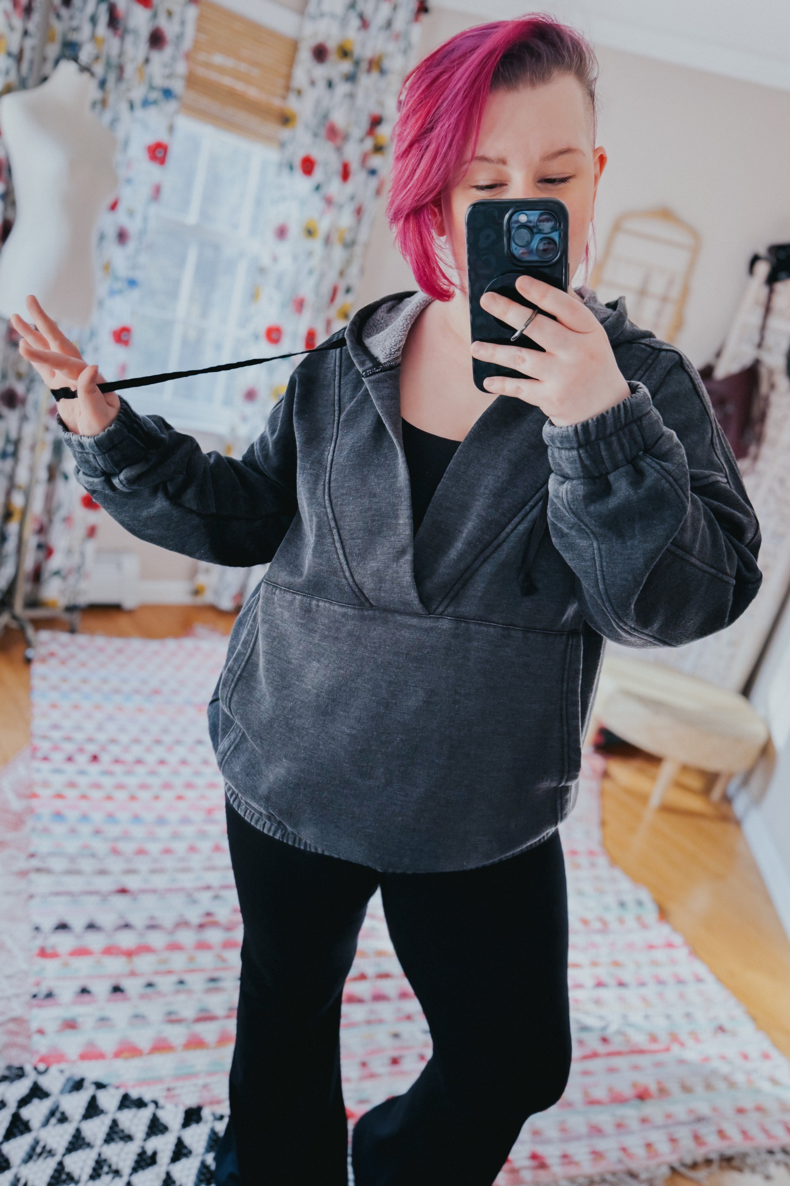 Woman with pink hair taking mirror selfie wearing charcoal acid wash hoodie with elastic cuffs and black pants indoors