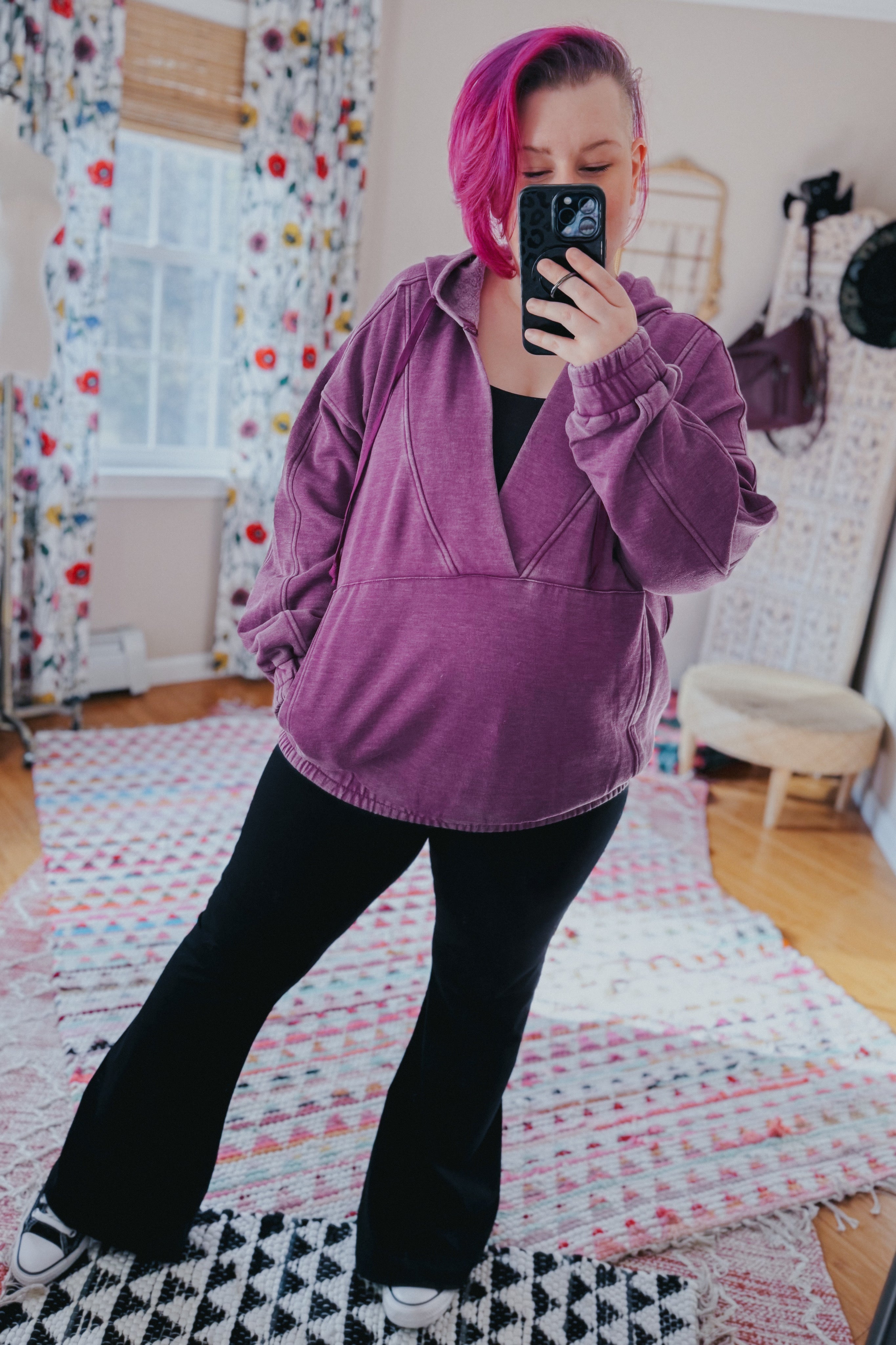 Woman with magenta hair wearing plum acid wash hoodie paired with black flared pants taking selfie in cozy room