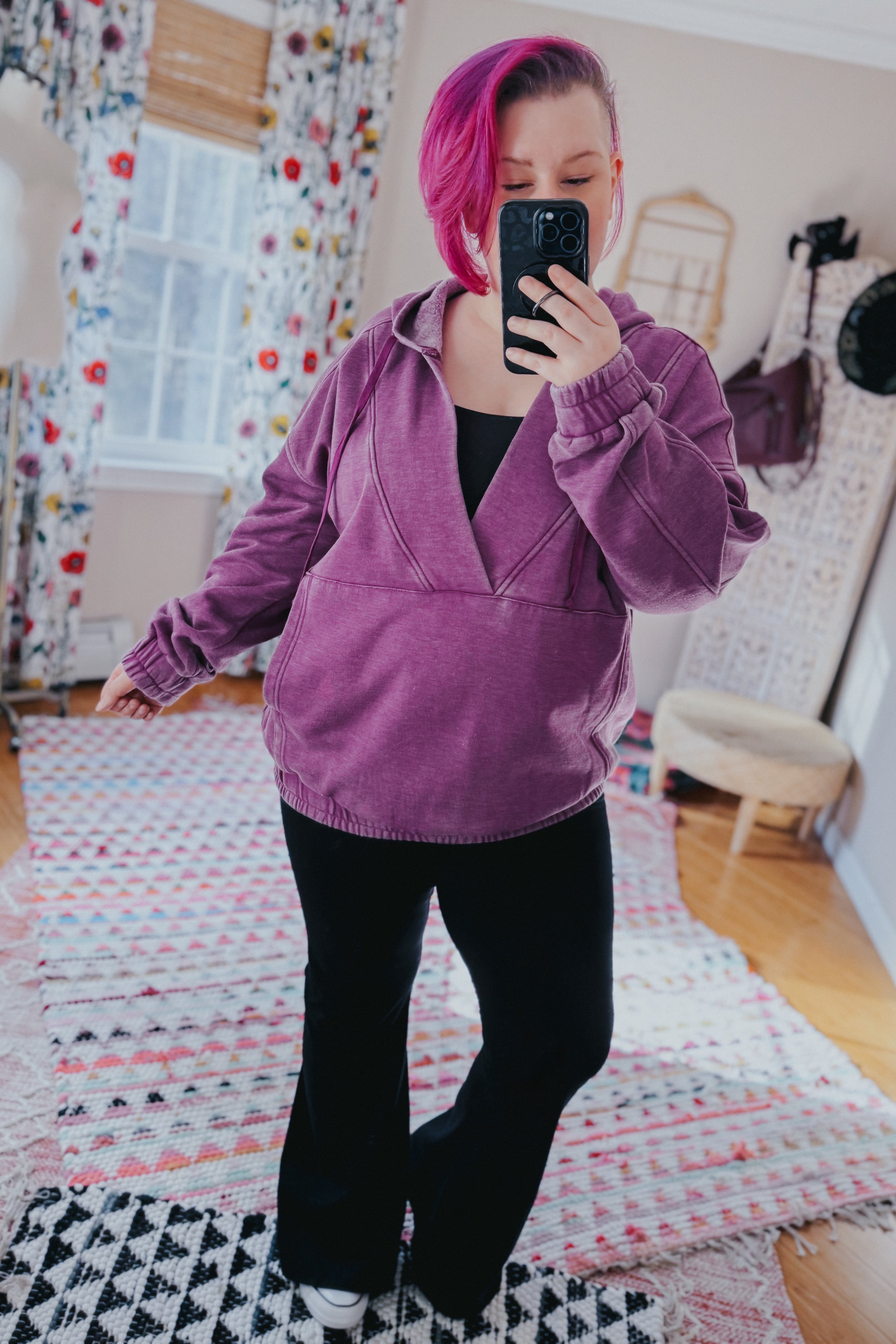 Woman with pink hair wearing plum acid wash hoodie with gathered cuffs and black pants taking indoor mirror selfie
