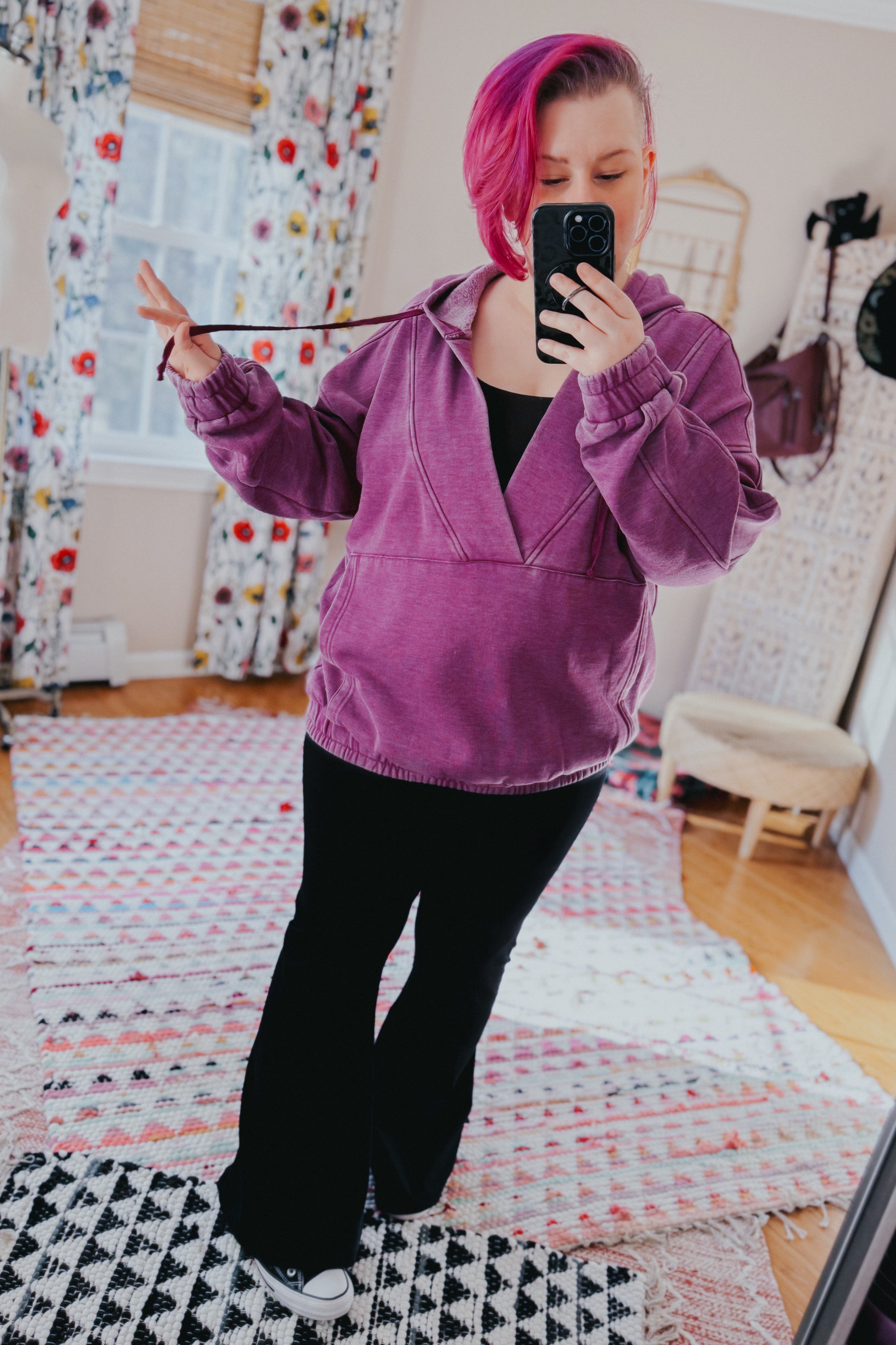 Woman with magenta hair wearing plum acid wash hoodie with drawstrings and black pants taking mirror selfie indoors