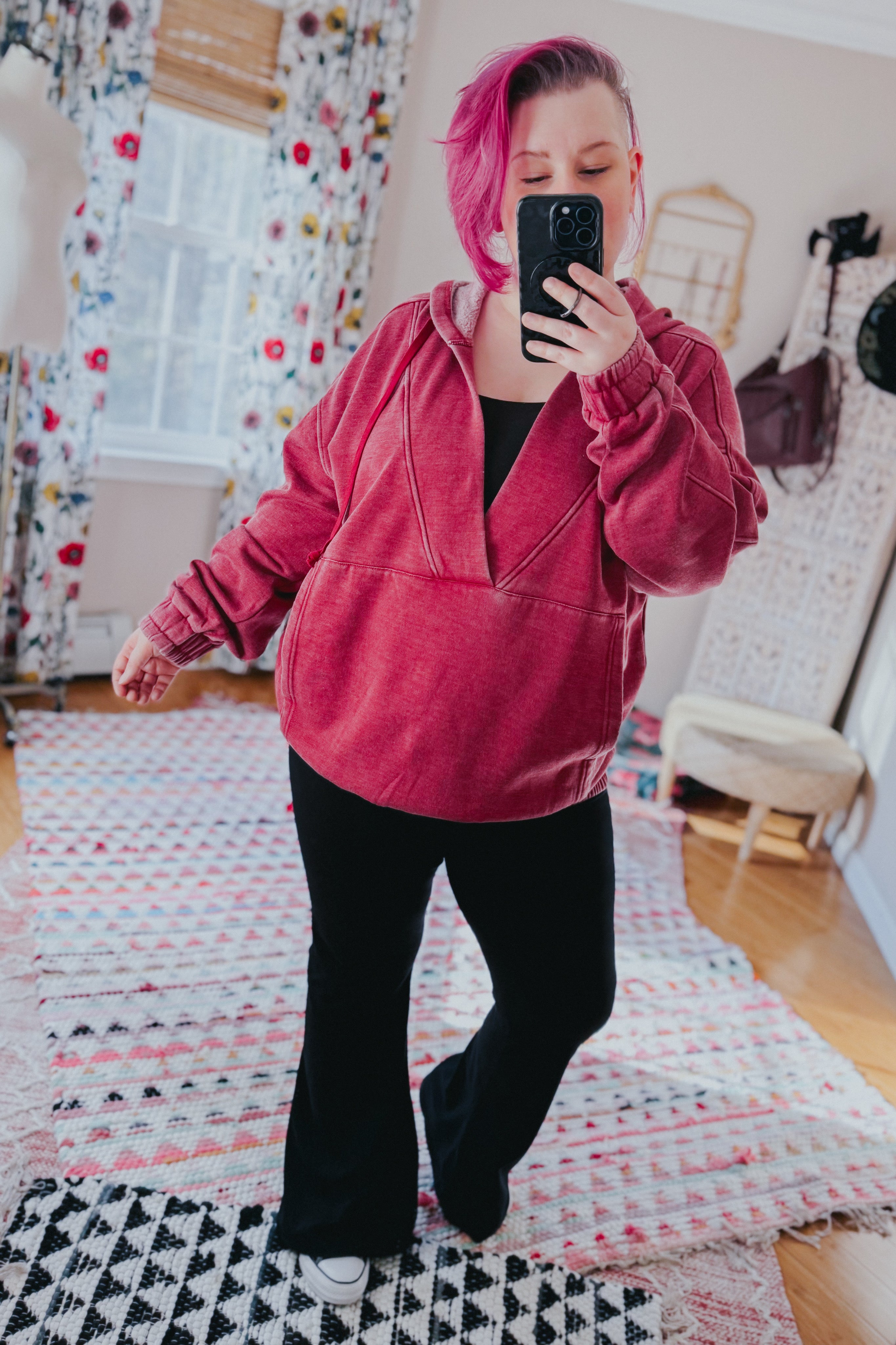 Woman with magenta hair wearing wine acid wash hoodie with gathered sleeves and black pants taking indoor mirror selfie