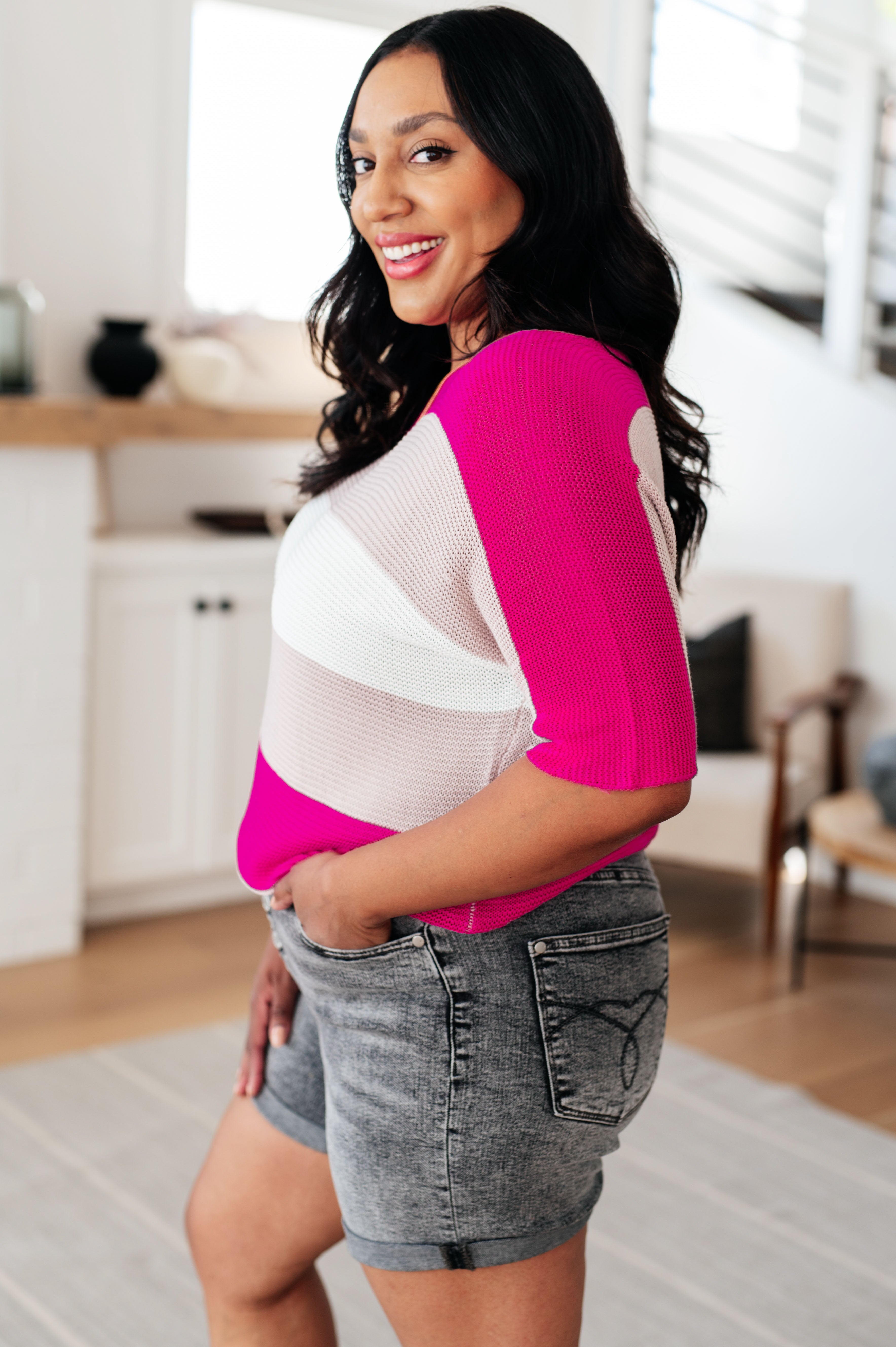 Side view of woman wearing a pink and white color-blocked short sleeve knit top with rolled denim shorts indoors