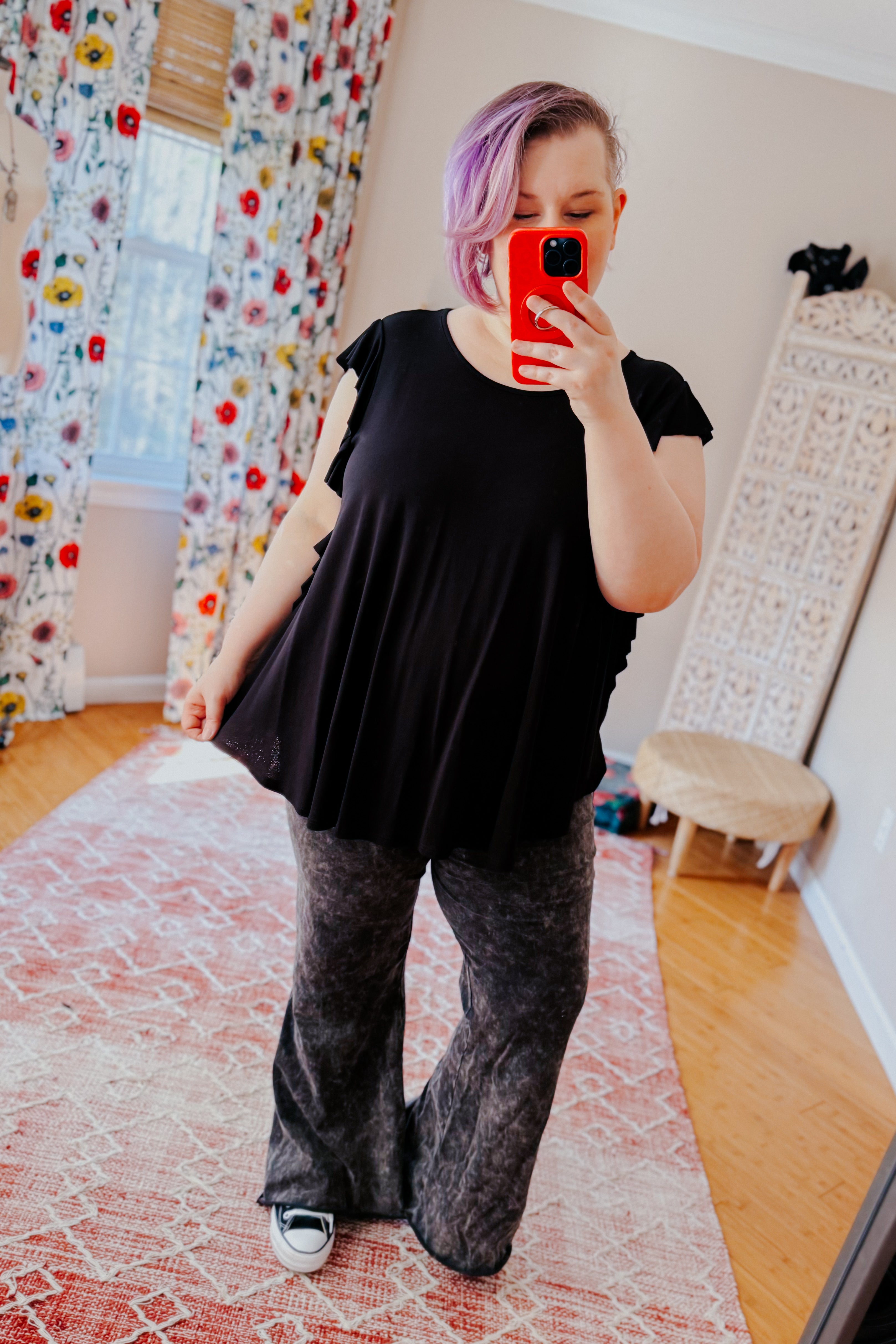 Woman with lavender hair wearing black ruffle sleeve top and patterned wide-leg pants taking mirror selfie in bright room