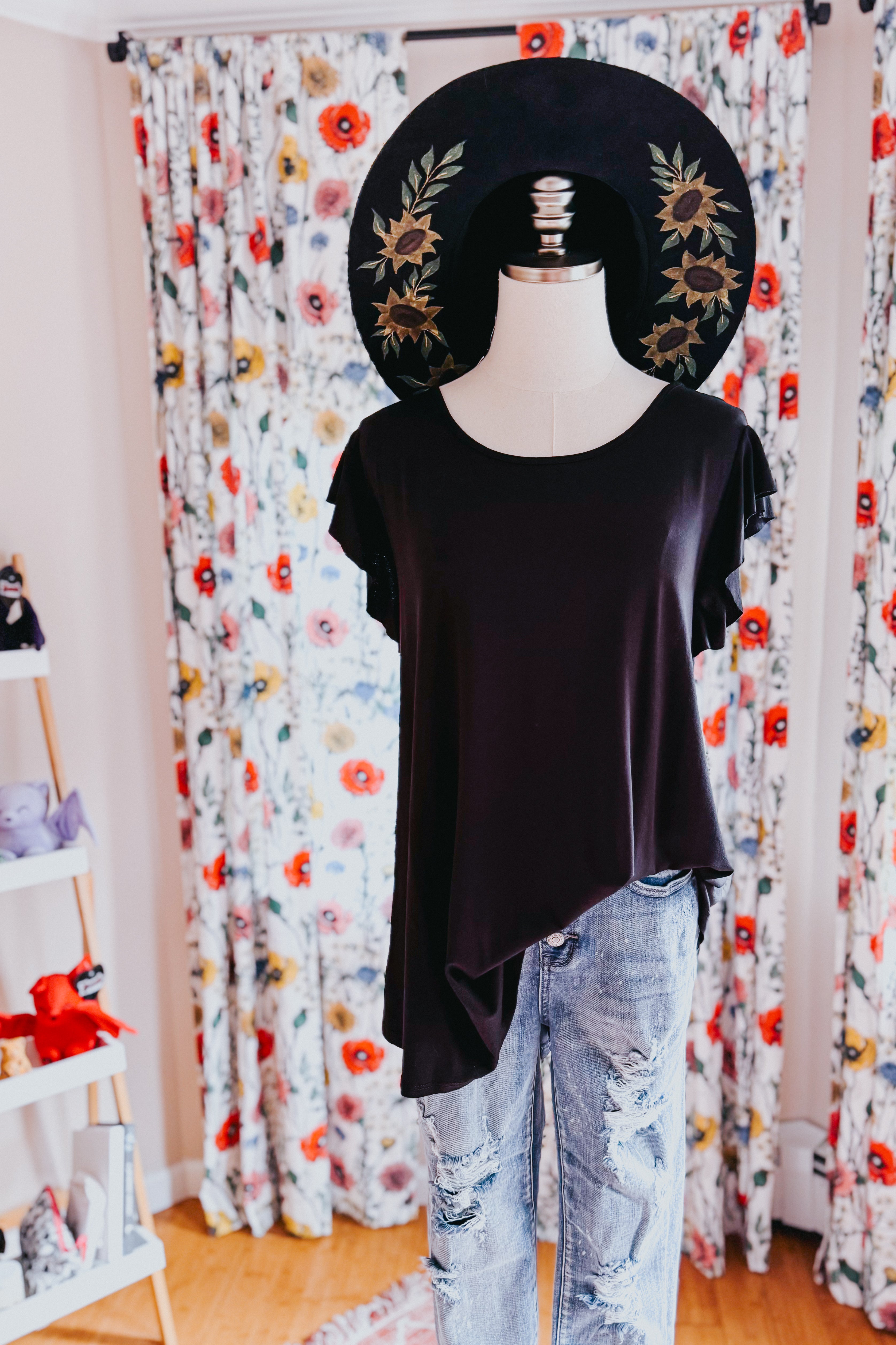 Black asymmetrical ruffle sleeve top paired with distressed light-wash jeans and wide-brimmed floral embroidered hat on mannequin