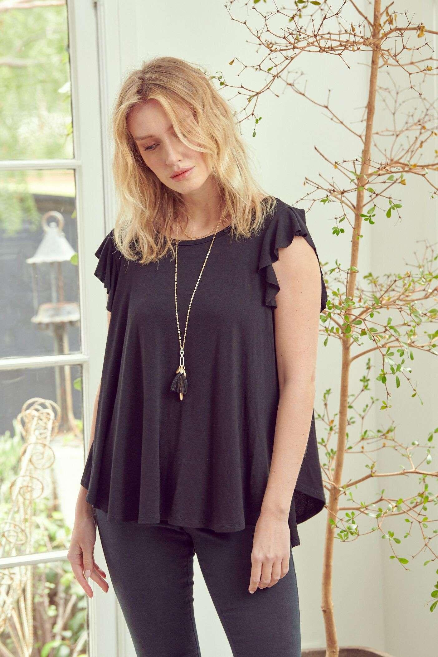 Woman wearing black sleeveless top with ruffle sleeves and long pendant necklace standing by window and plant