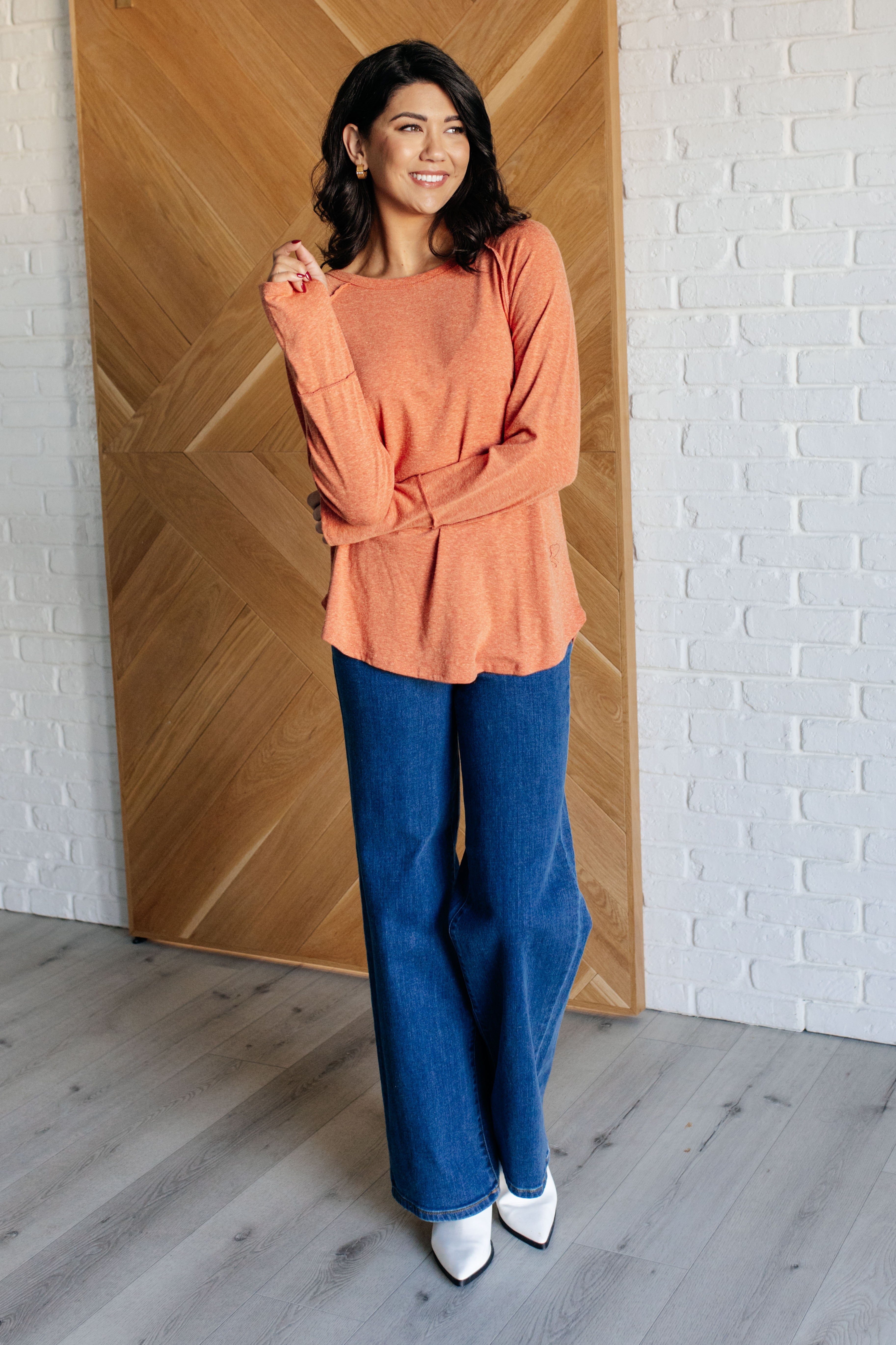Smiling woman wearing orange long sleeve top and wide-leg jeans standing indoors with wooden door background
