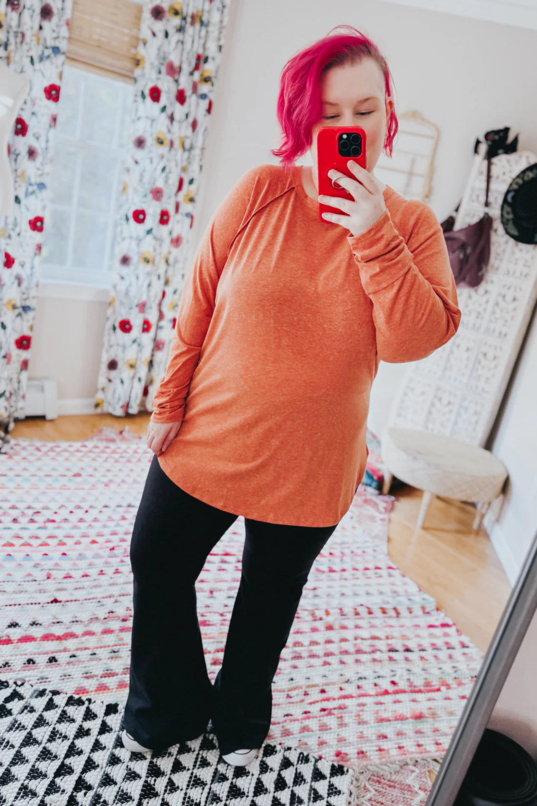 Woman with vibrant pink hair wearing orange long sleeve top and black pants taking mirror selfie indoors