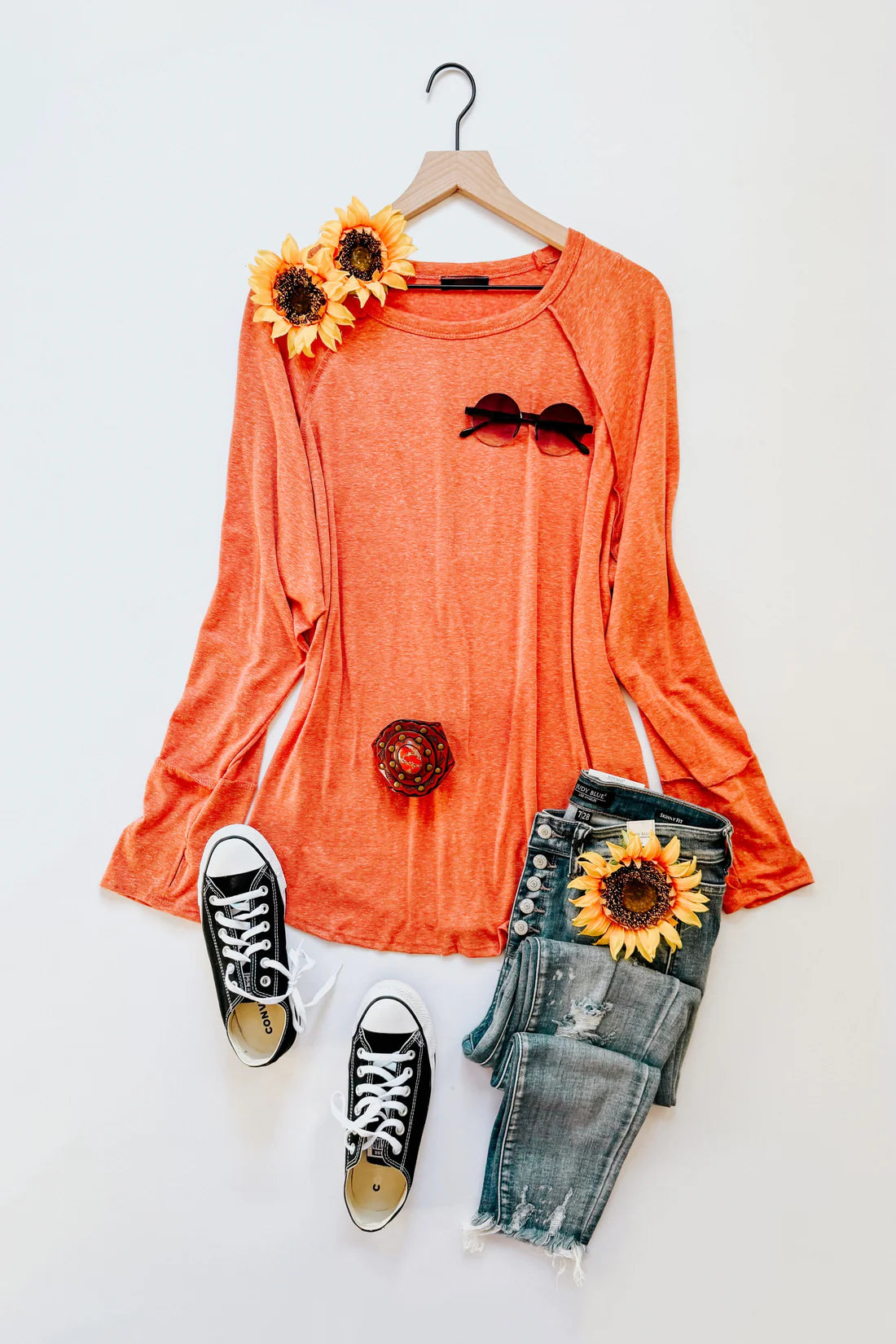 Orange long sleeve top paired with distressed jeans, black sneakers, and sunflower accents laid flat with sunglasses and ring accessory