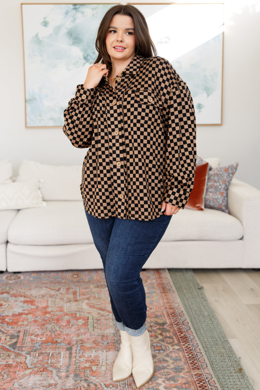 Plus-size model wearing brown checkered shacket with dark jeans and white boots indoors on patterned rugs
