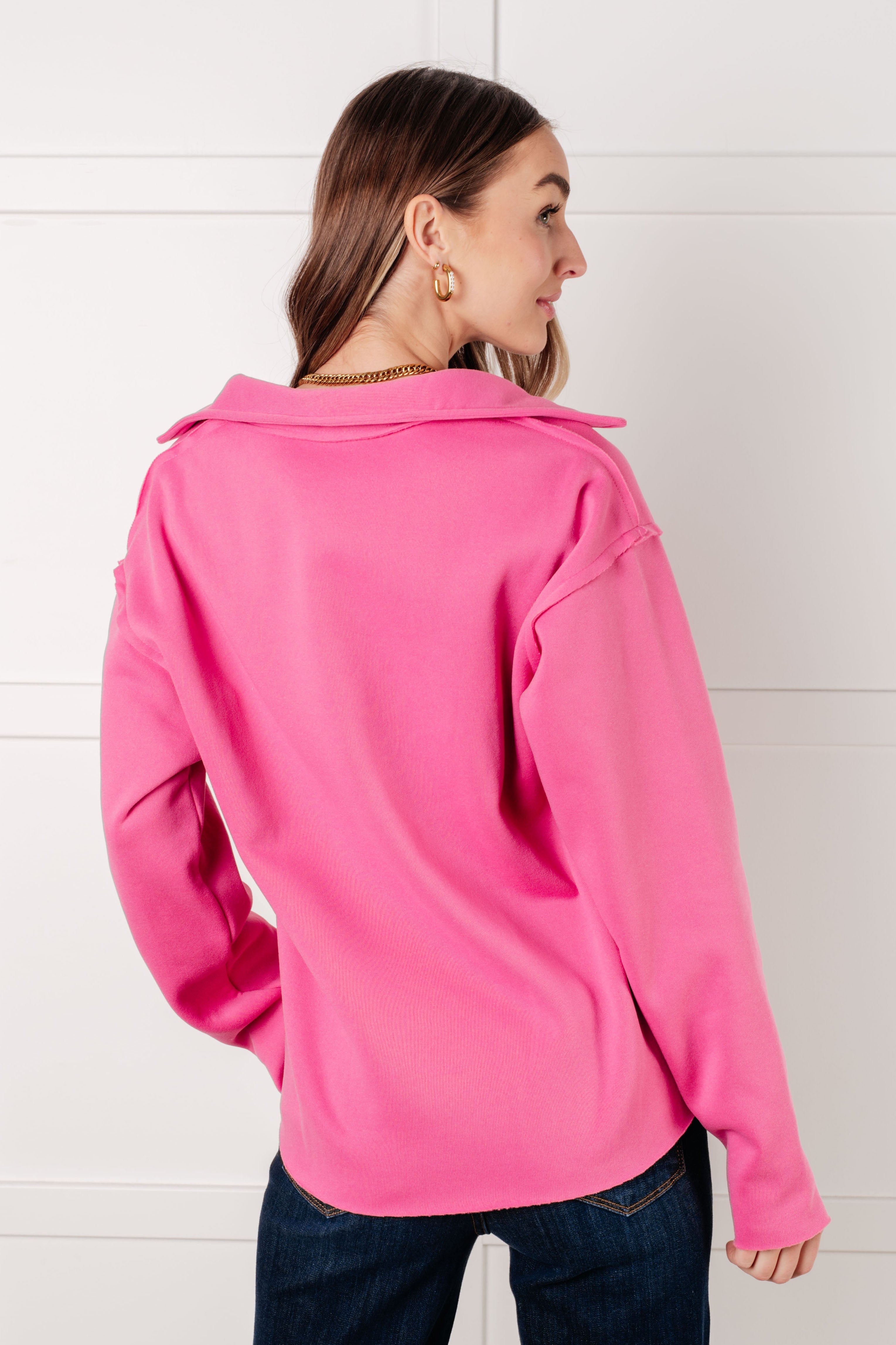 Woman wearing a hot pink colored pullover with exposed seams and dark blue jeans with a white background.