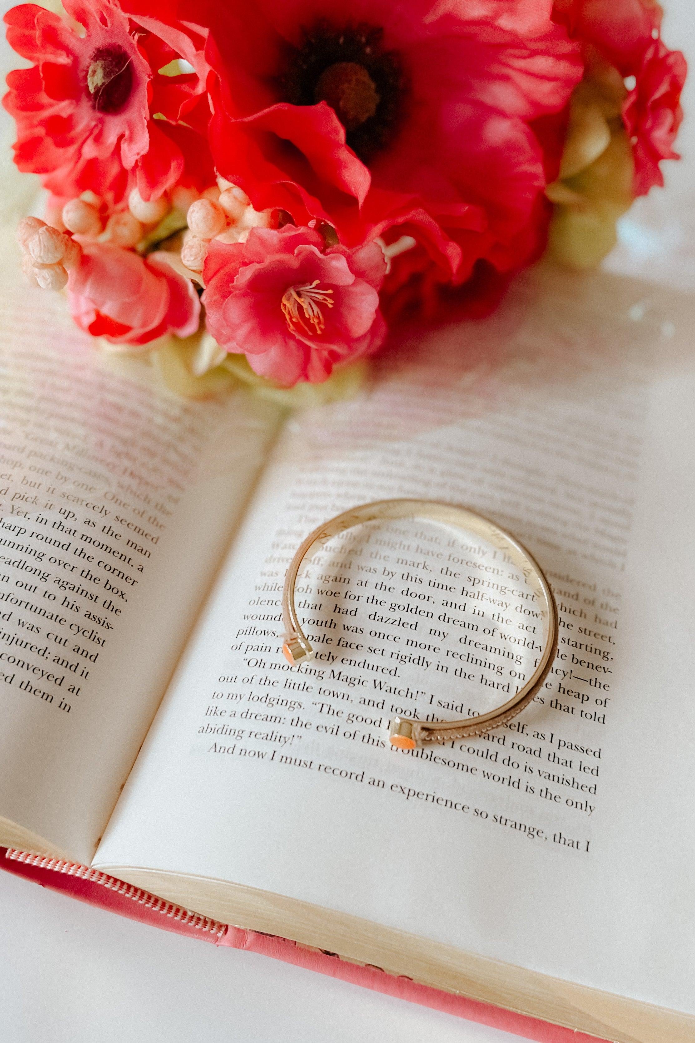Beige stackable bracelet resting on open book near bright red and pink floral bouquet with soft natural light