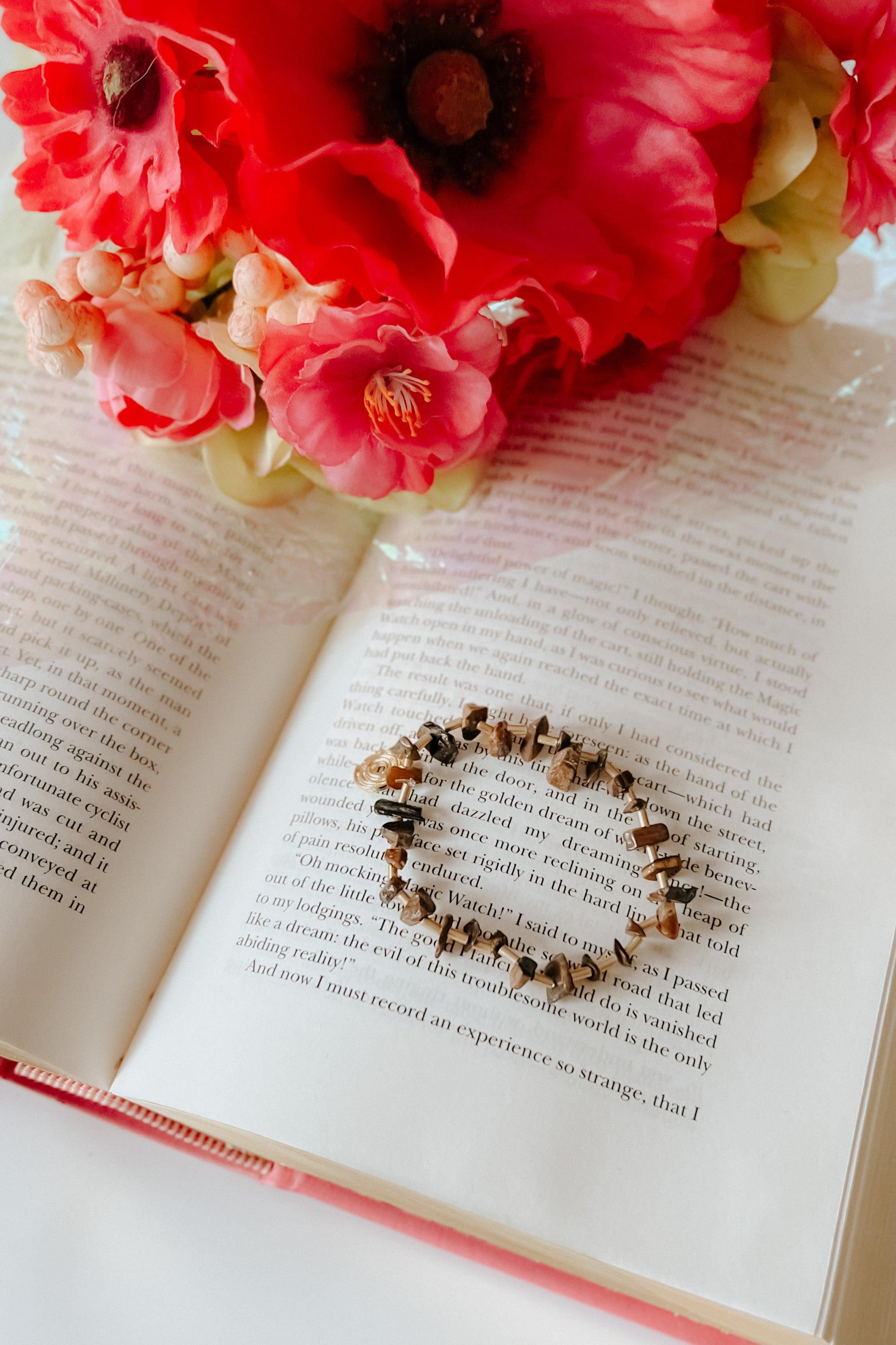 Beige stackable bracelet with geometric beads resting on an open book next to bright red and pink flowers