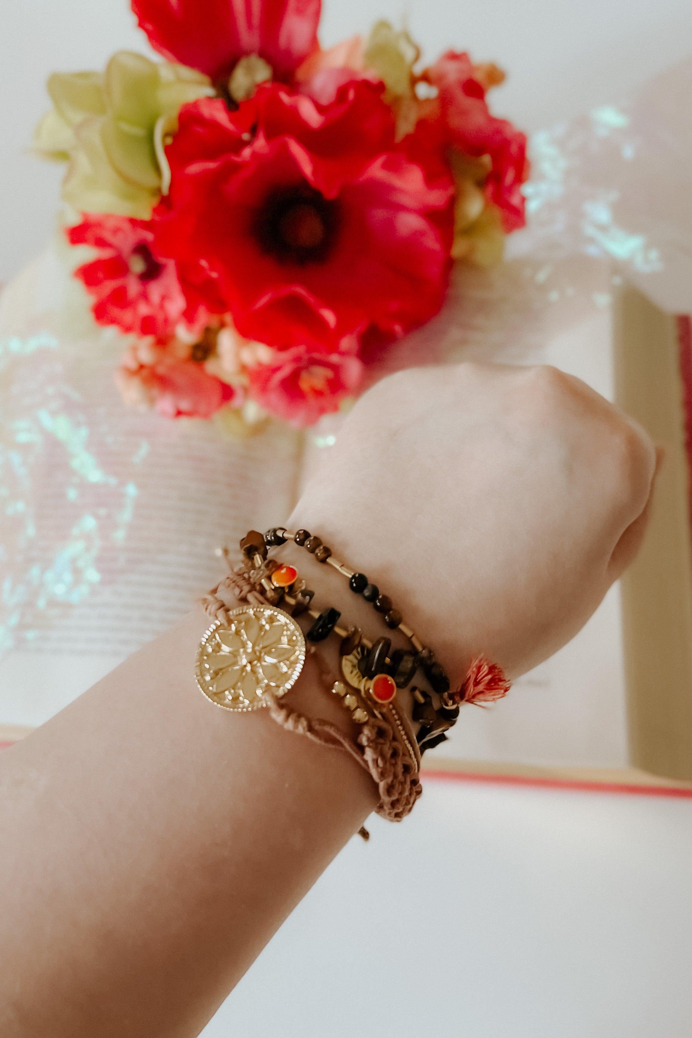 Beige stackable bracelets with gold charm and red accents worn on wrist above open book and red flowers
