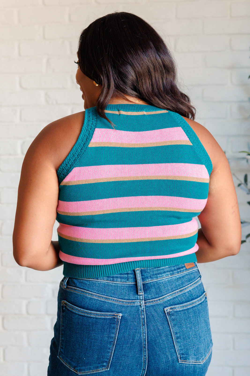 Back view of woman wearing blue knit striped tank with pink and brown stripes paired with blue jeans indoors