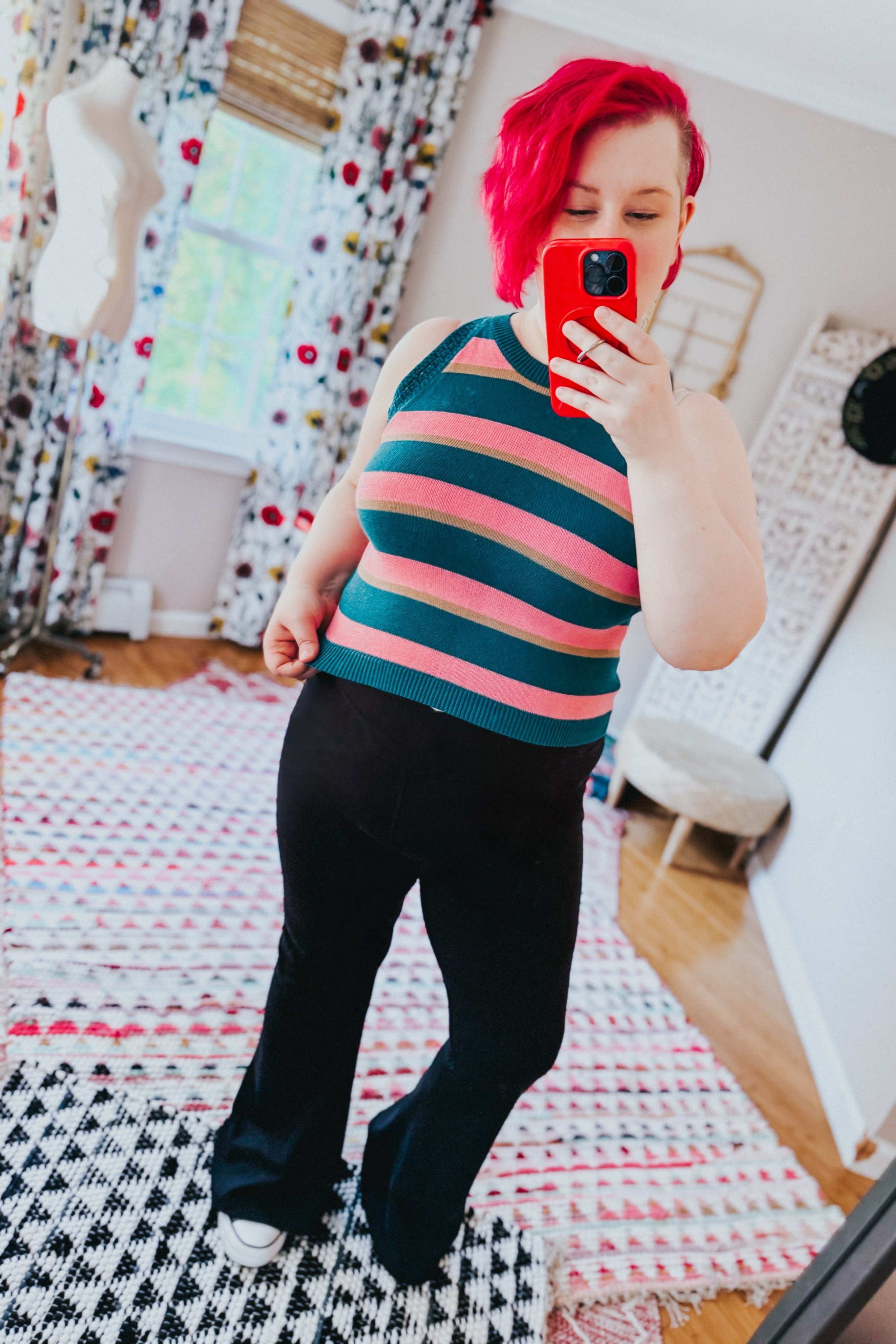 Woman with red hair wearing a blue and pink striped knit tank top paired with black pants standing indoors