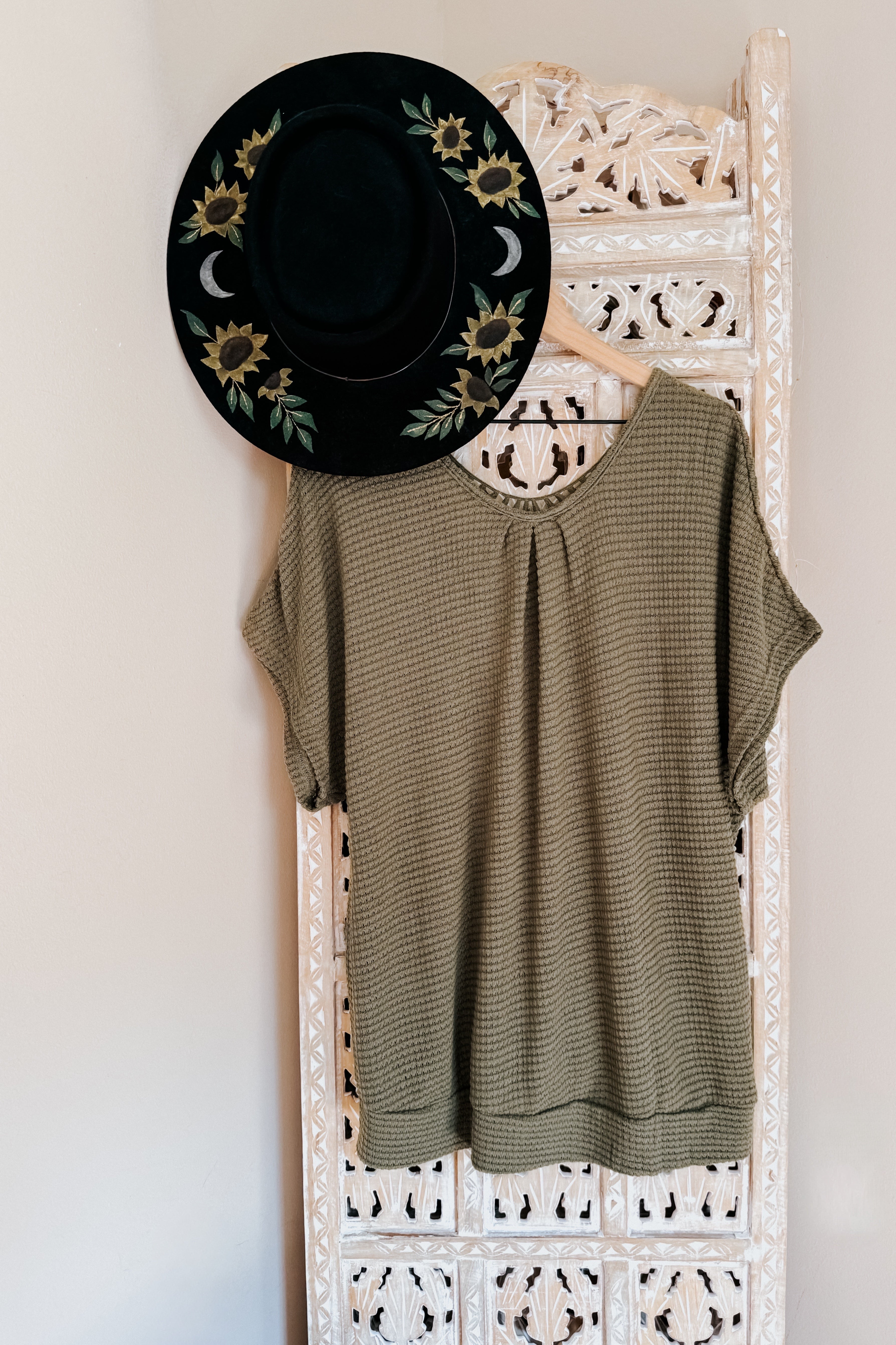 Olive waffle knit loose top with short sleeves hanging on intricate white carved screen next to black floral hat
