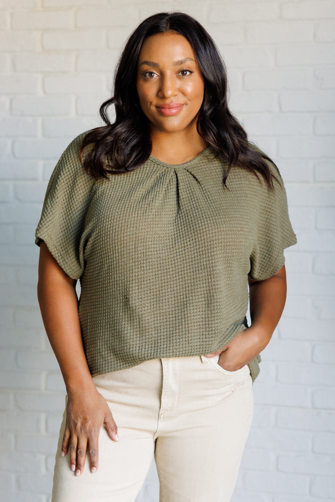 Woman wearing olive textured short sleeve top with pleated neckline paired with light beige pants indoors