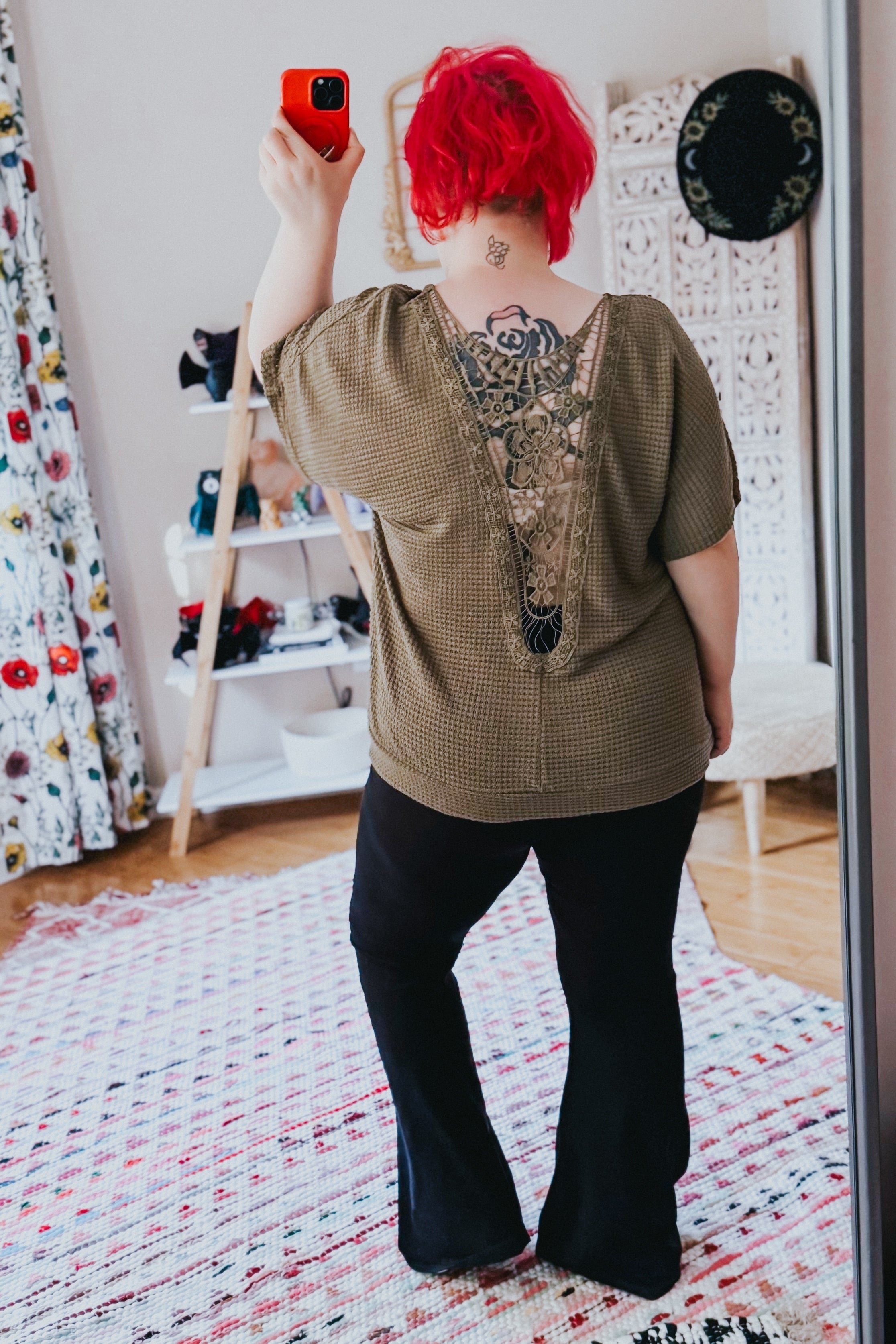 Woman with bright red hair wearing olive top with lace back detail paired with black pants taking a mirror selfie indoors