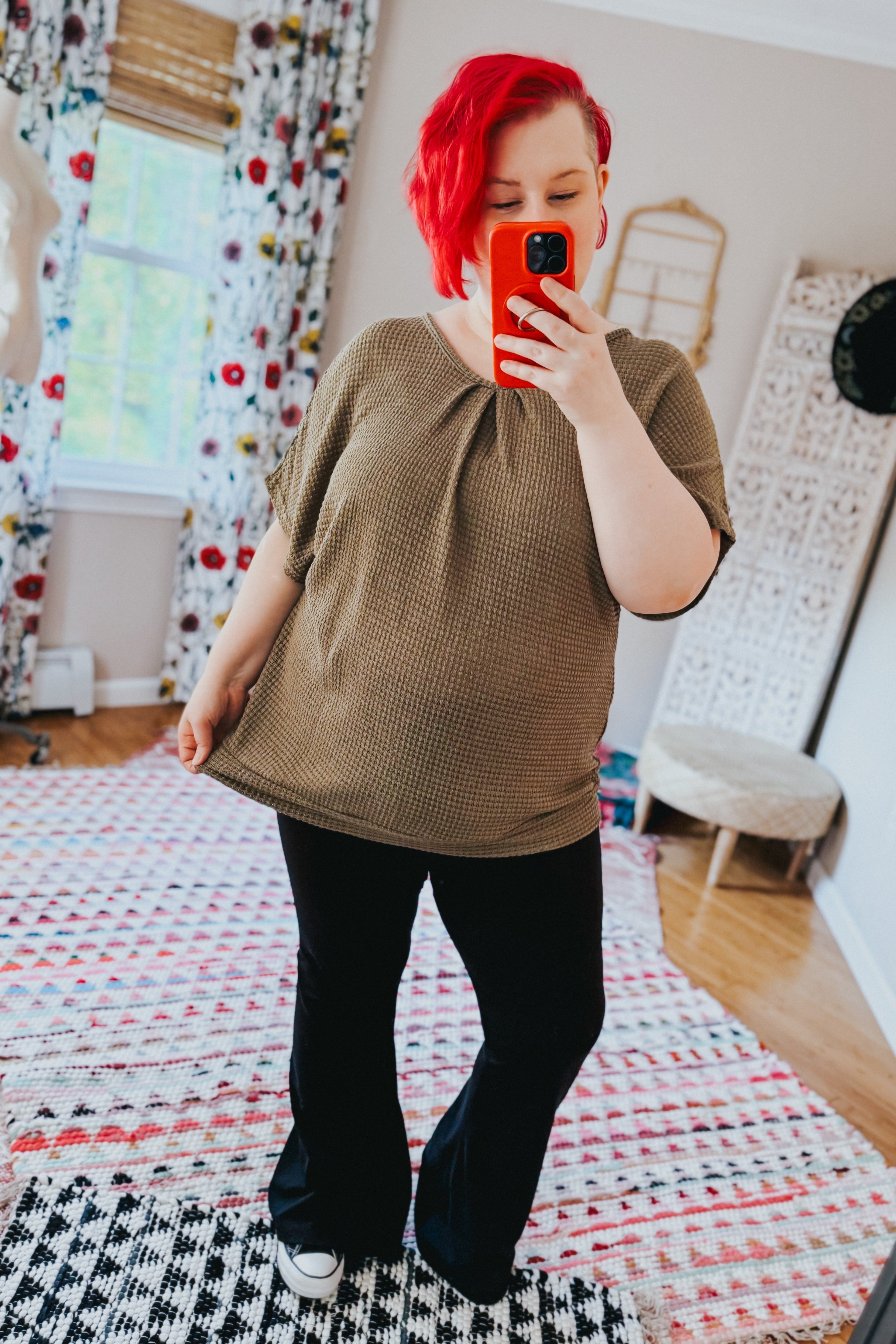 Woman with red hair wearing olive textured top with pleated neckline paired with black flared pants and white sneakers indoors