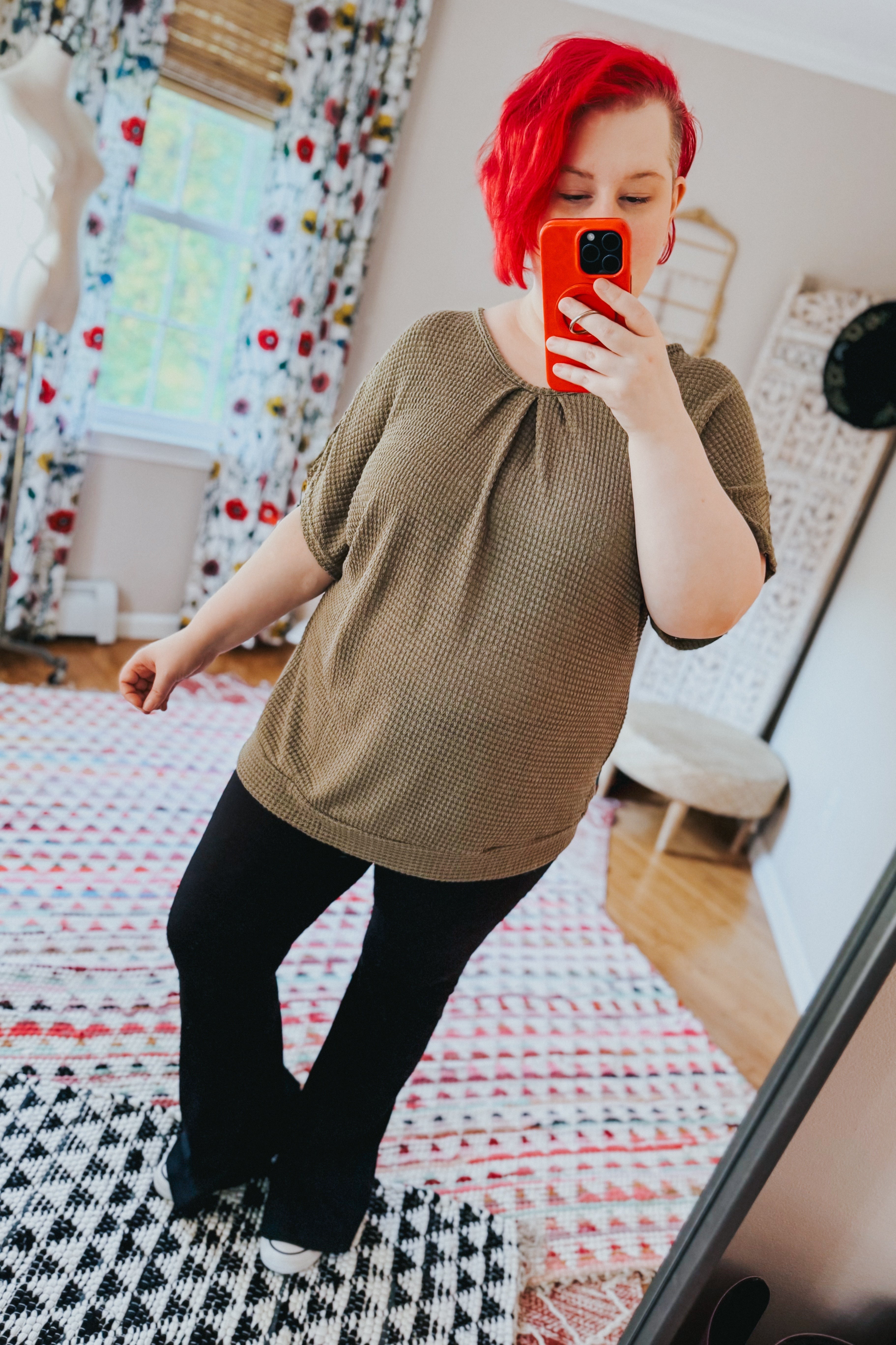 Woman with red hair wearing olive textured short sleeve top paired with black pants taking a mirror selfie indoors