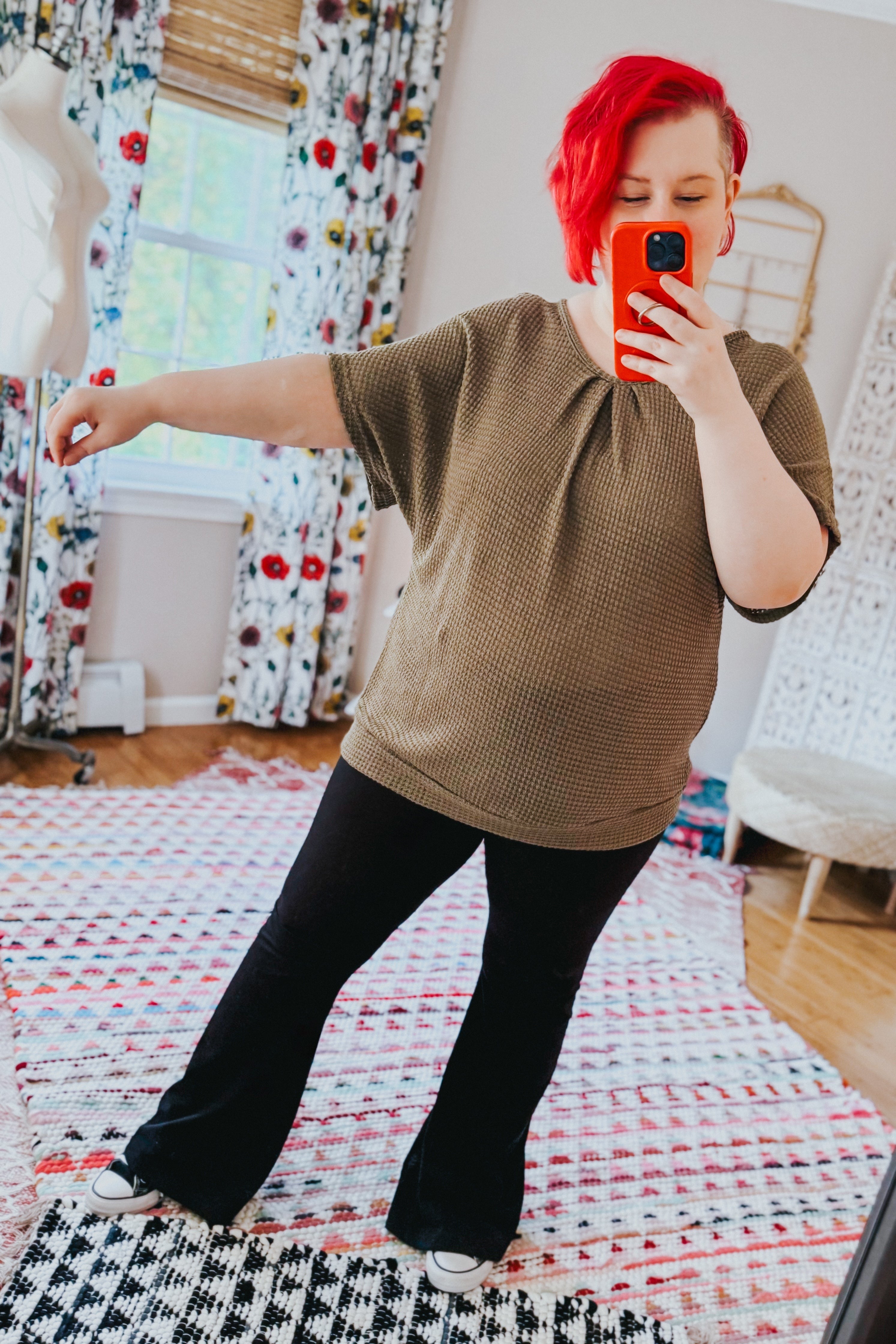 Woman with bright red hair wearing olive textured top and black pants taking mirror selfie in cozy bright room