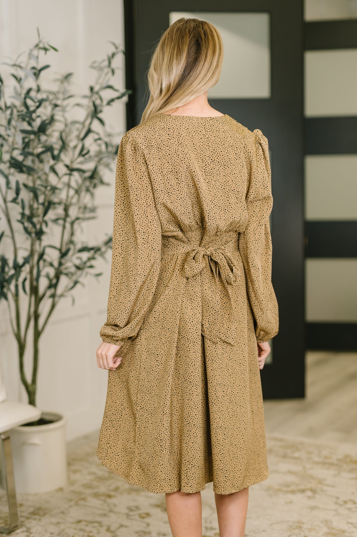 Back view of a woman wearing a camel long-sleeve dress with a tied waist and softly flared skirt indoors