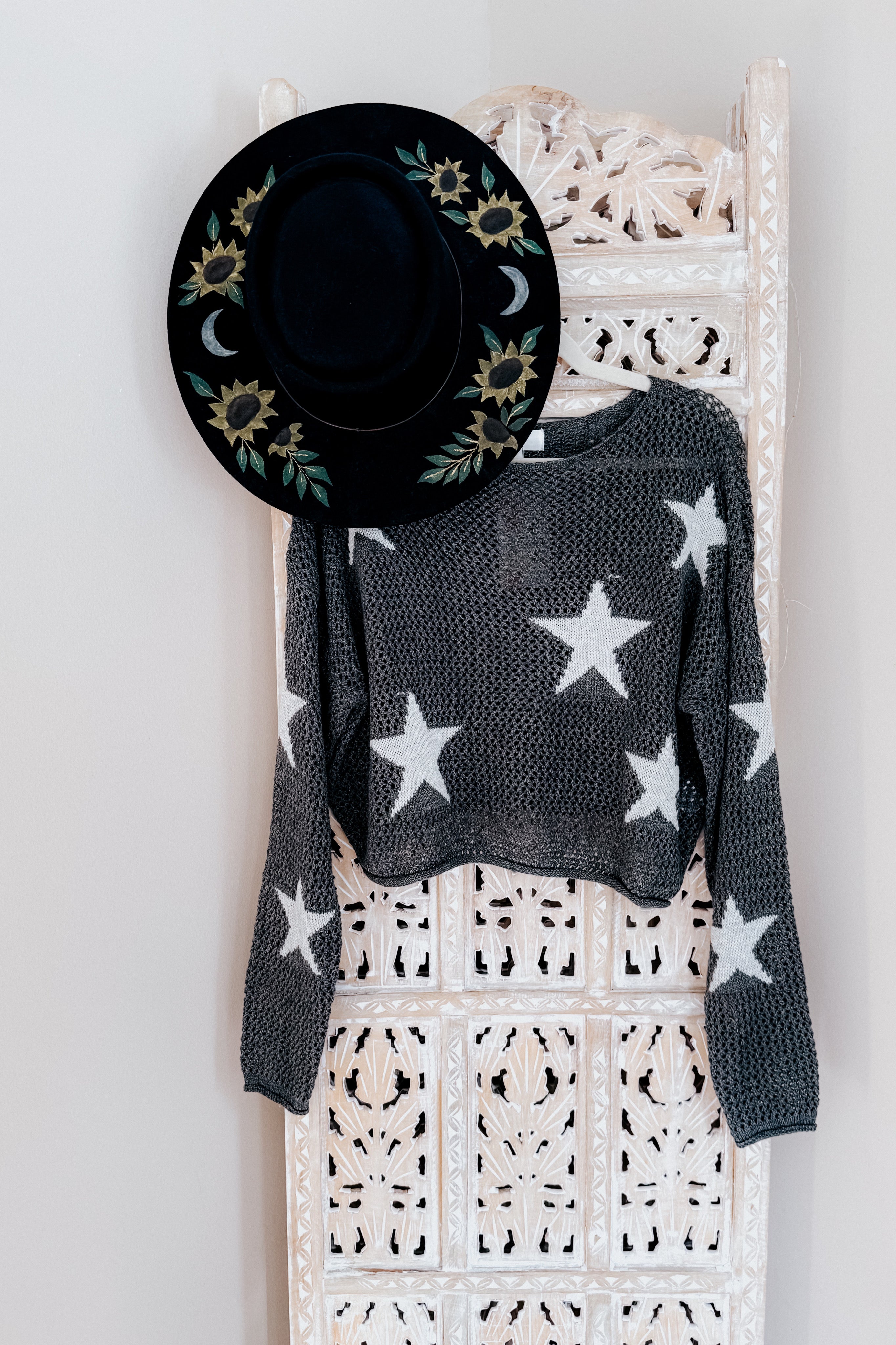 Charcoal mesh sweater with white stars hanging on ornate white wooden panel paired with black floral embroidered hat