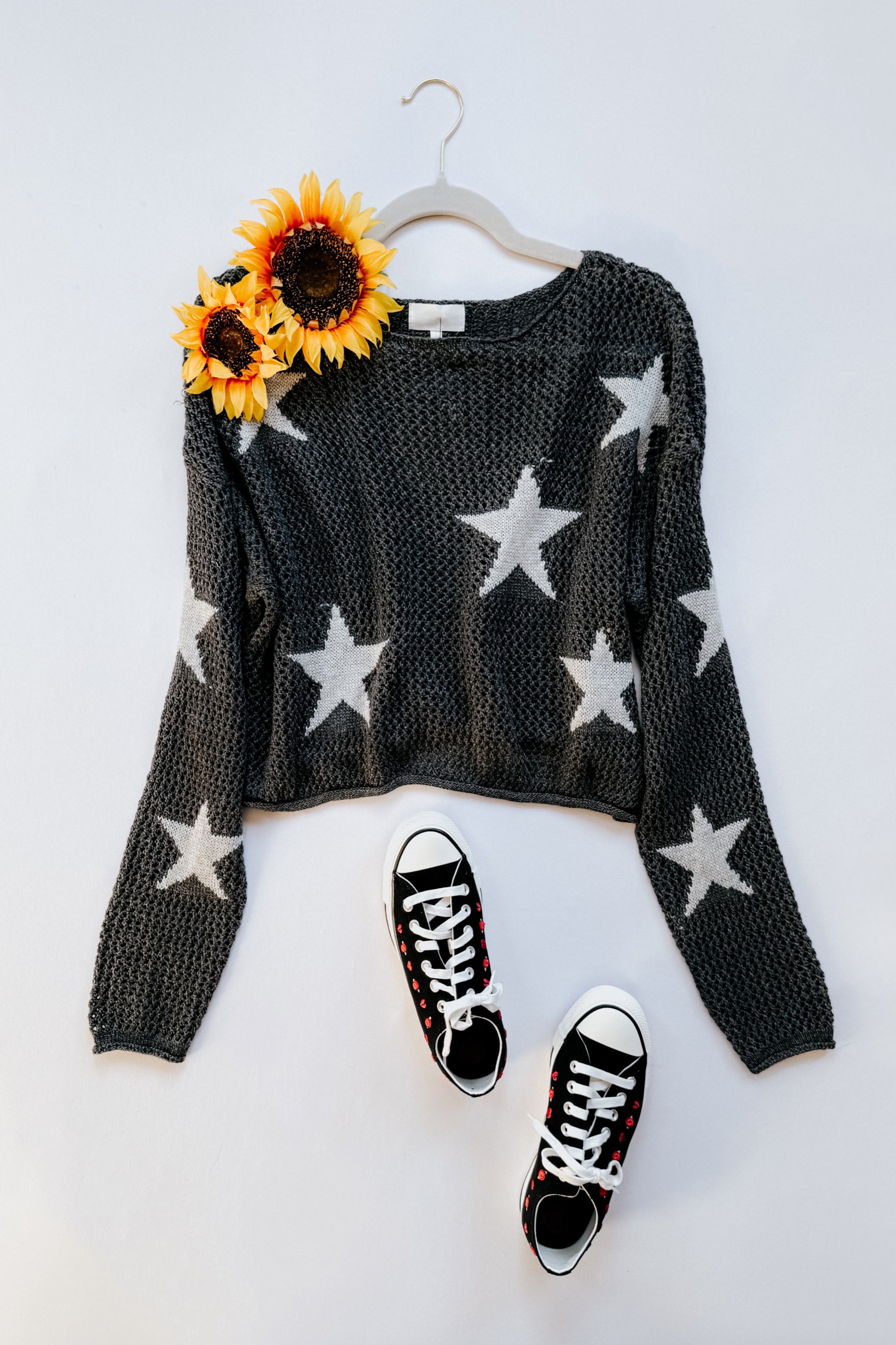 Charcoal mesh sweater with white stars paired with black sneakers and bright sunflowers on hanger against white background