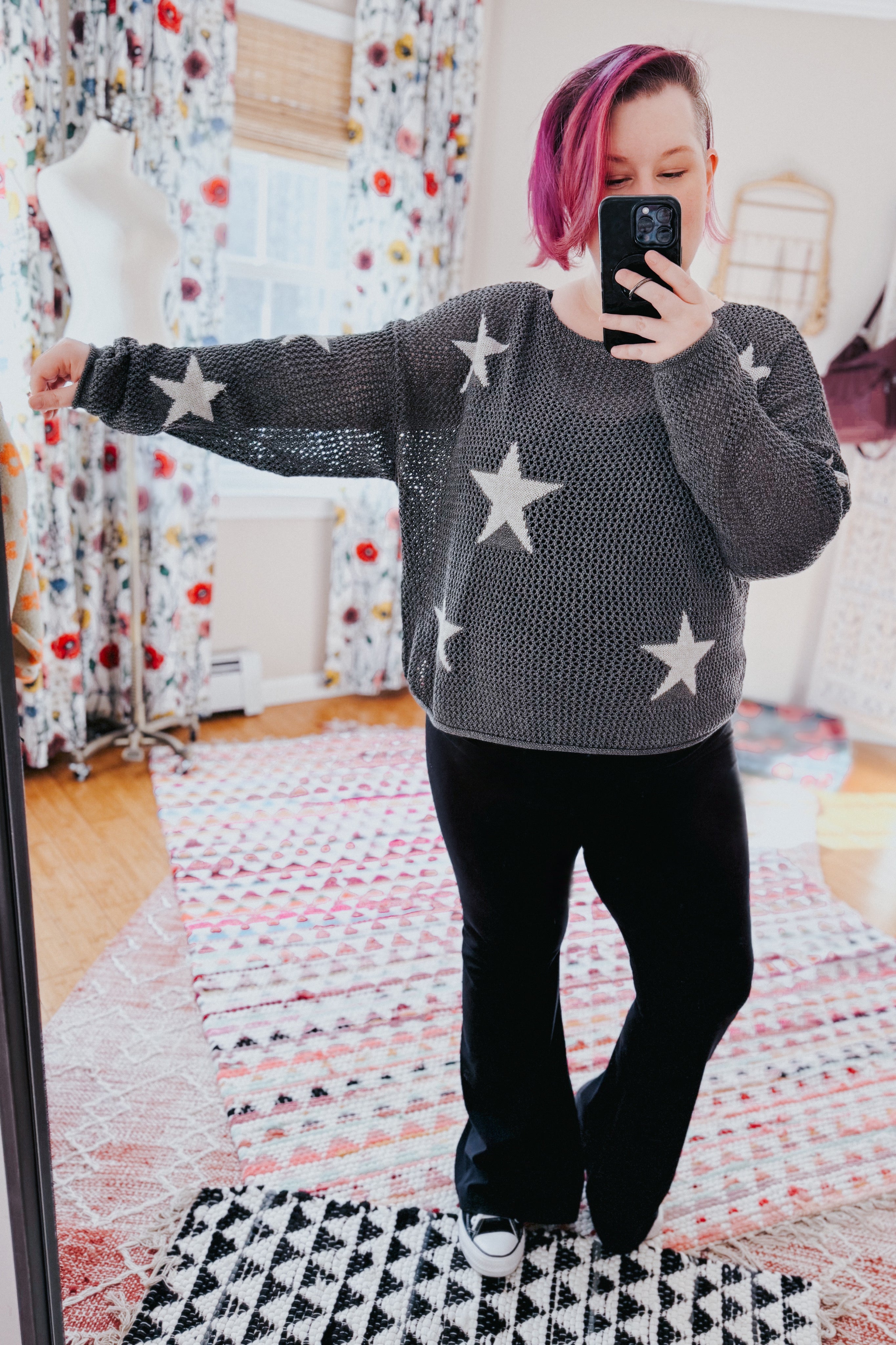 Woman with pink hair wearing charcoal mesh sweater top with star patterns paired with black pants taking a mirror selfie indoors