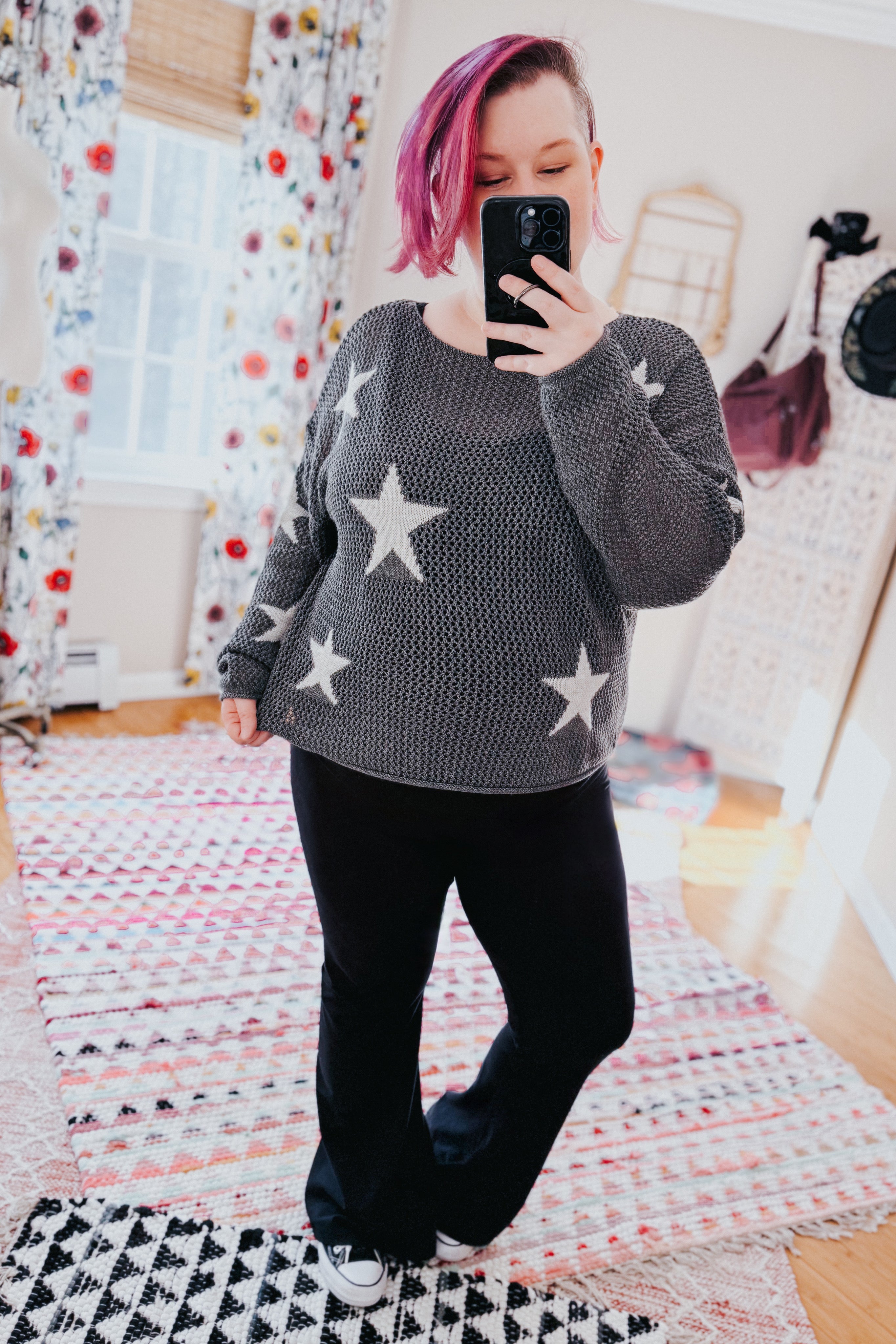 Woman with pink hair wearing charcoal mesh sweater top with star patterns paired with black pants taking indoor mirror selfie