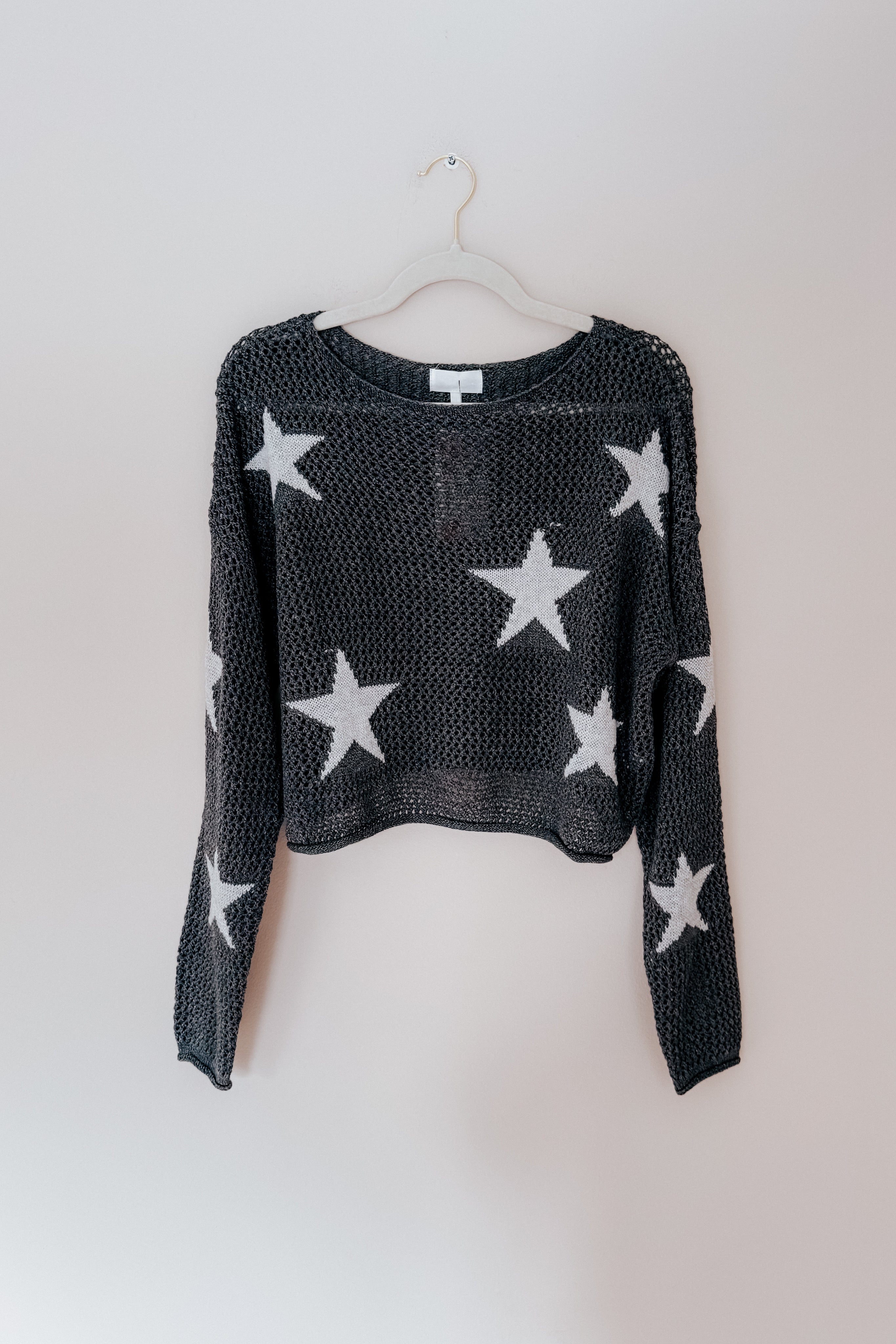 Charcoal mesh sweater with scattered white stars and relaxed fit hanging on light-colored hanger against plain wall