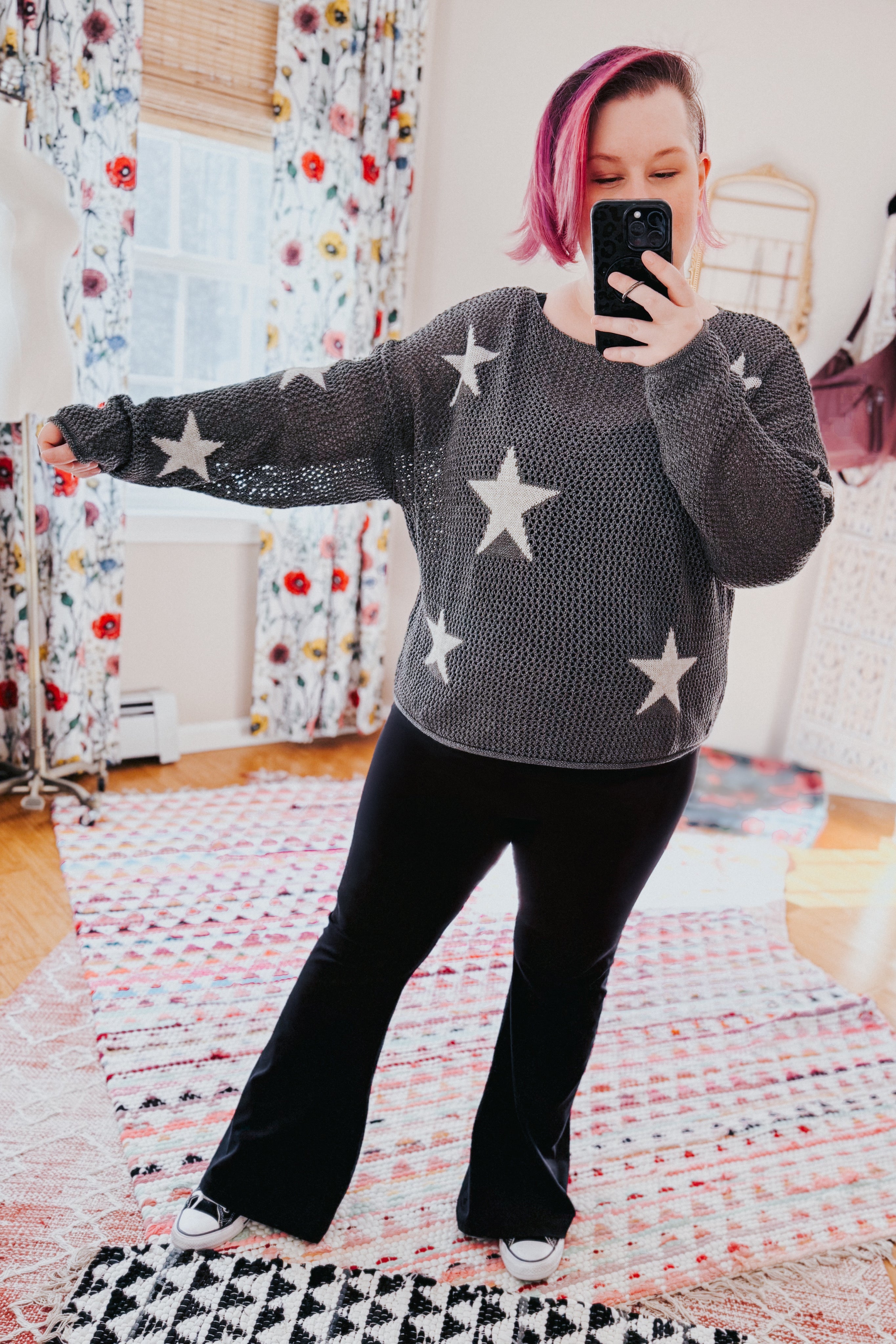 Woman with dyed pink hair wearing charcoal mesh sweater top with star patterns paired with black flared pants indoors