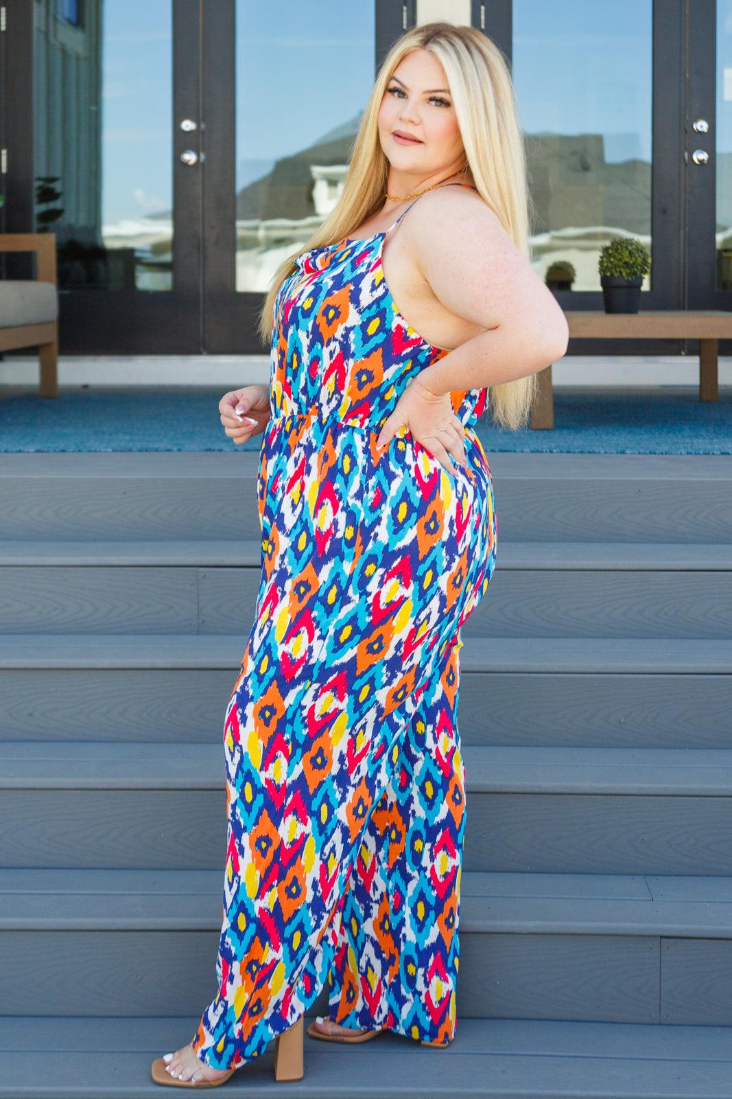 Plus-size model wearing a vibrant blue patterned jumpsuit standing sideways on outdoor wooden steps