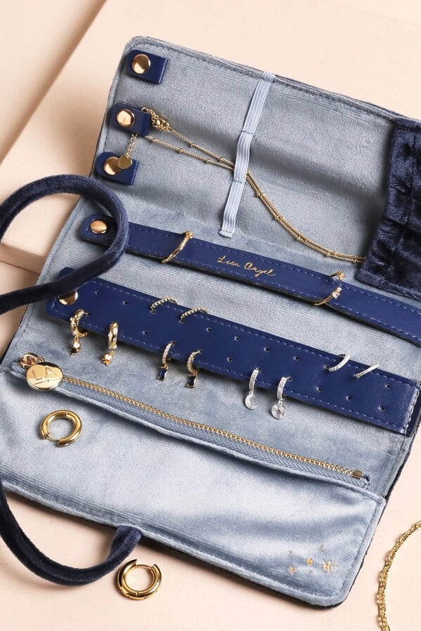 Open blue velvet jewelry roll with multiple gold and silver earrings and necklace inside and on beige surface