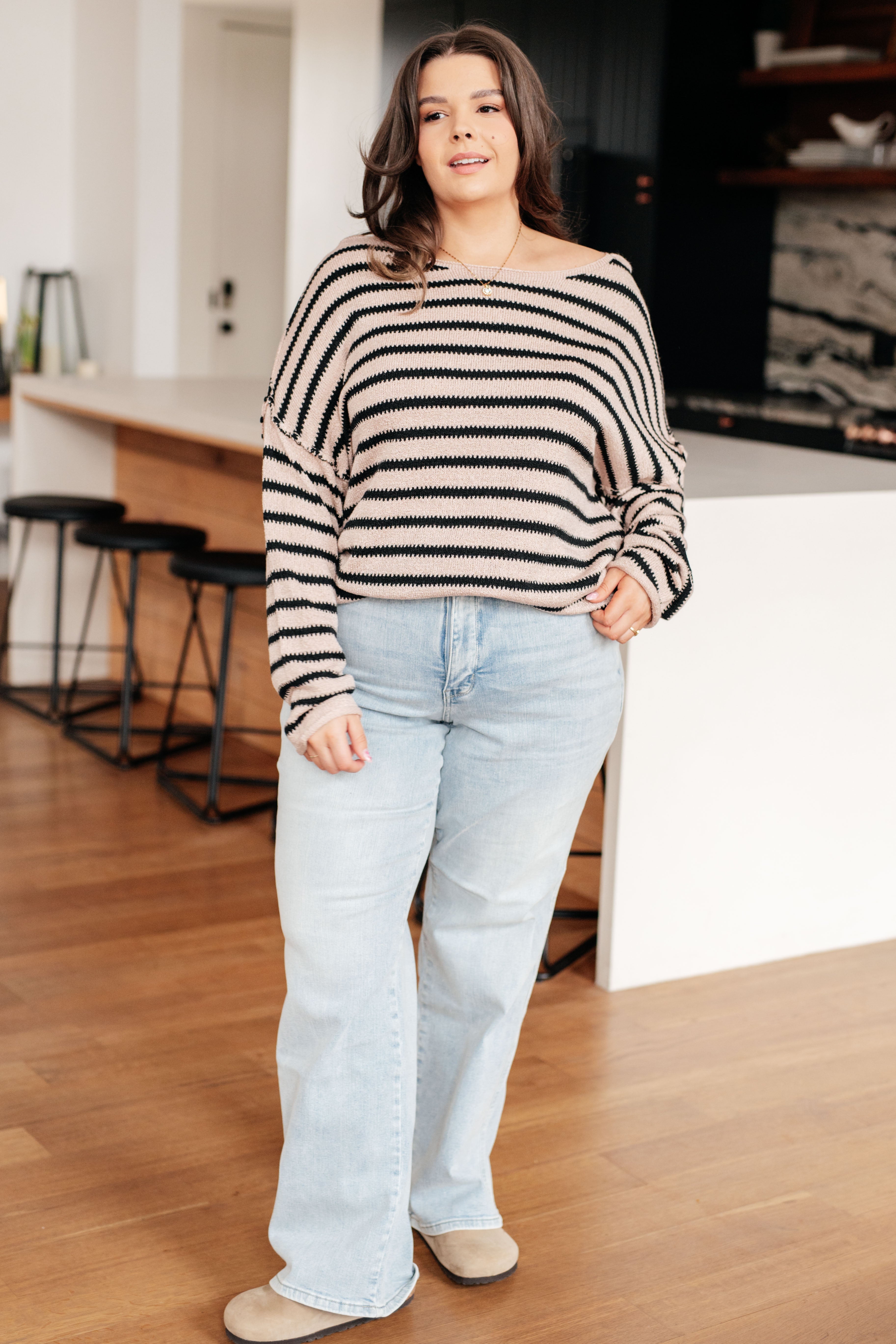 Woman wearing a taupe sweater with black stripes, light blue jeans and tan shoes indoors.