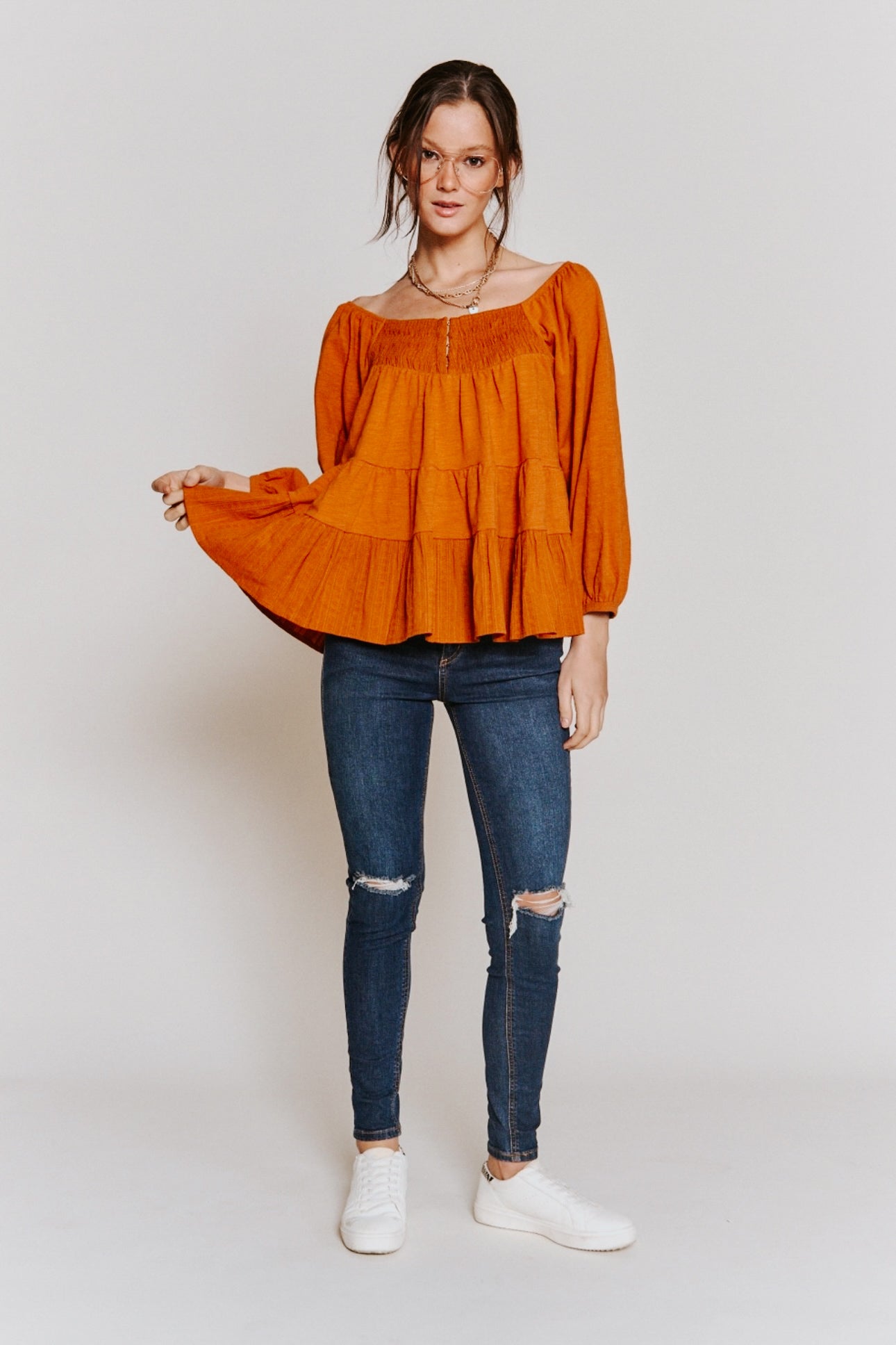 Woman wearing rust-colored ruched babydoll top styled with ripped skinny jeans and white sneakers on plain background