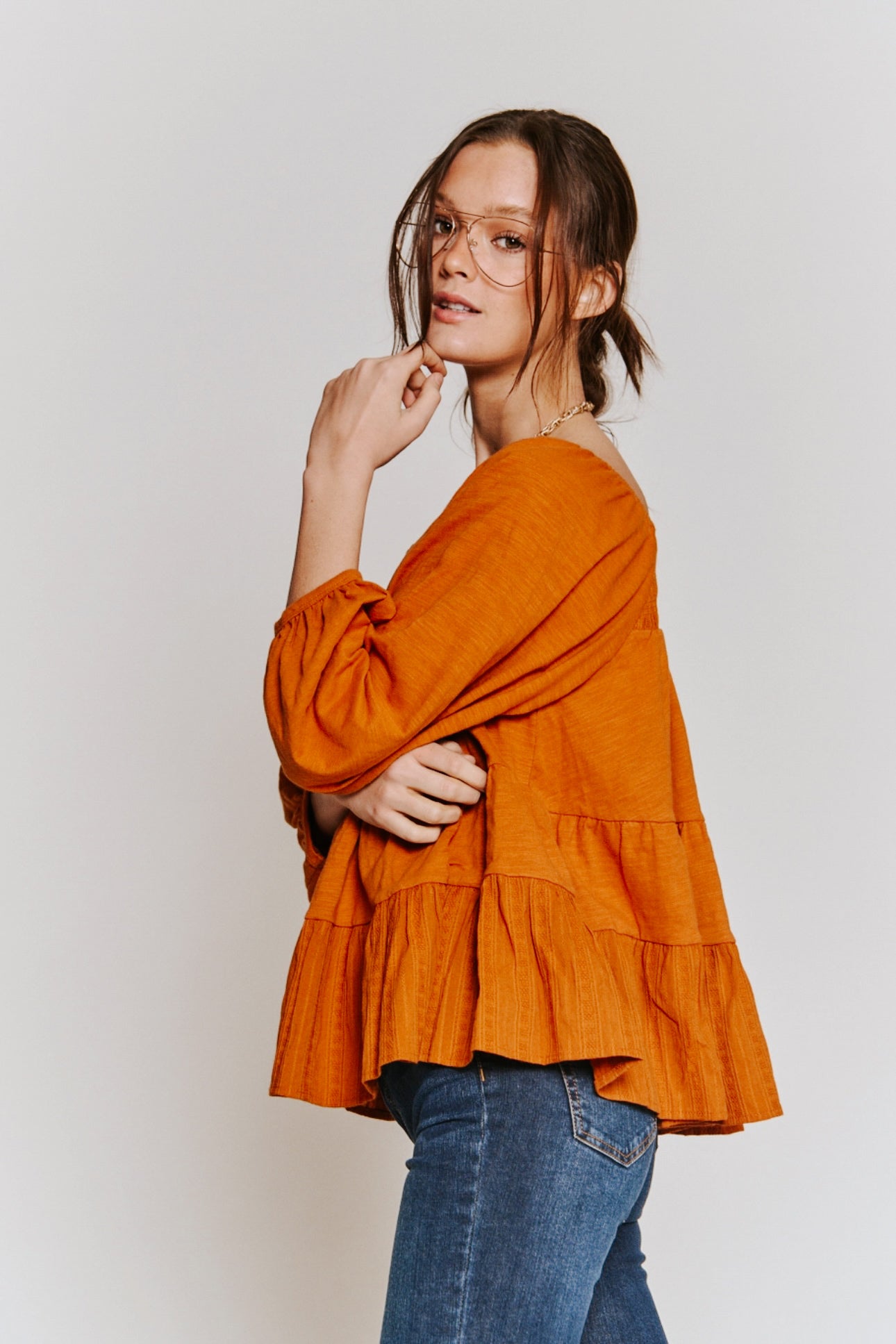 Woman wearing rust-colored ruched babydoll top paired with blue jeans and glasses against plain backdrop