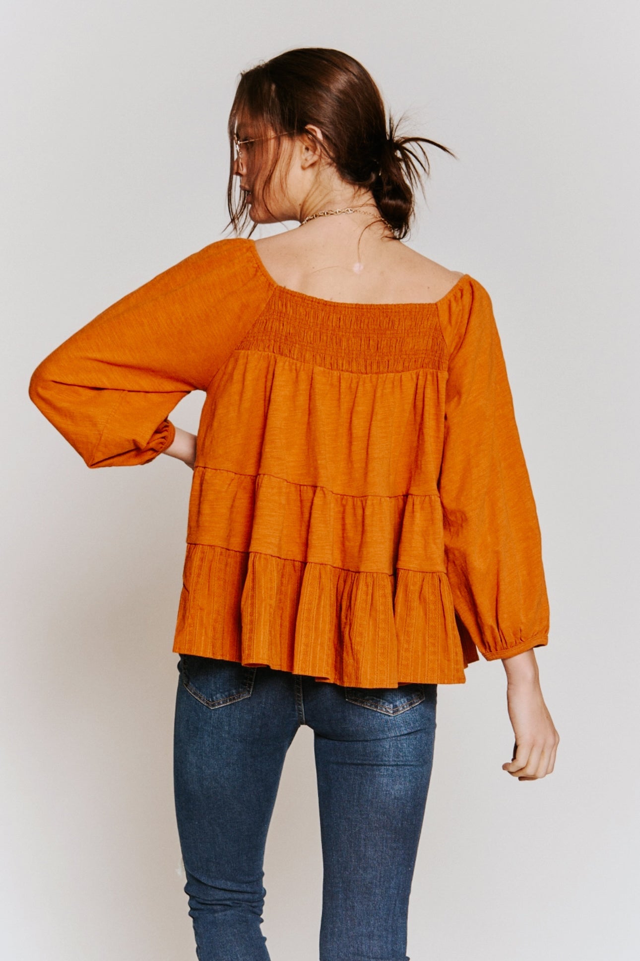 Woman wearing rust-colored ruched babydoll top with tiered back and puffed sleeves paired with blue jeans