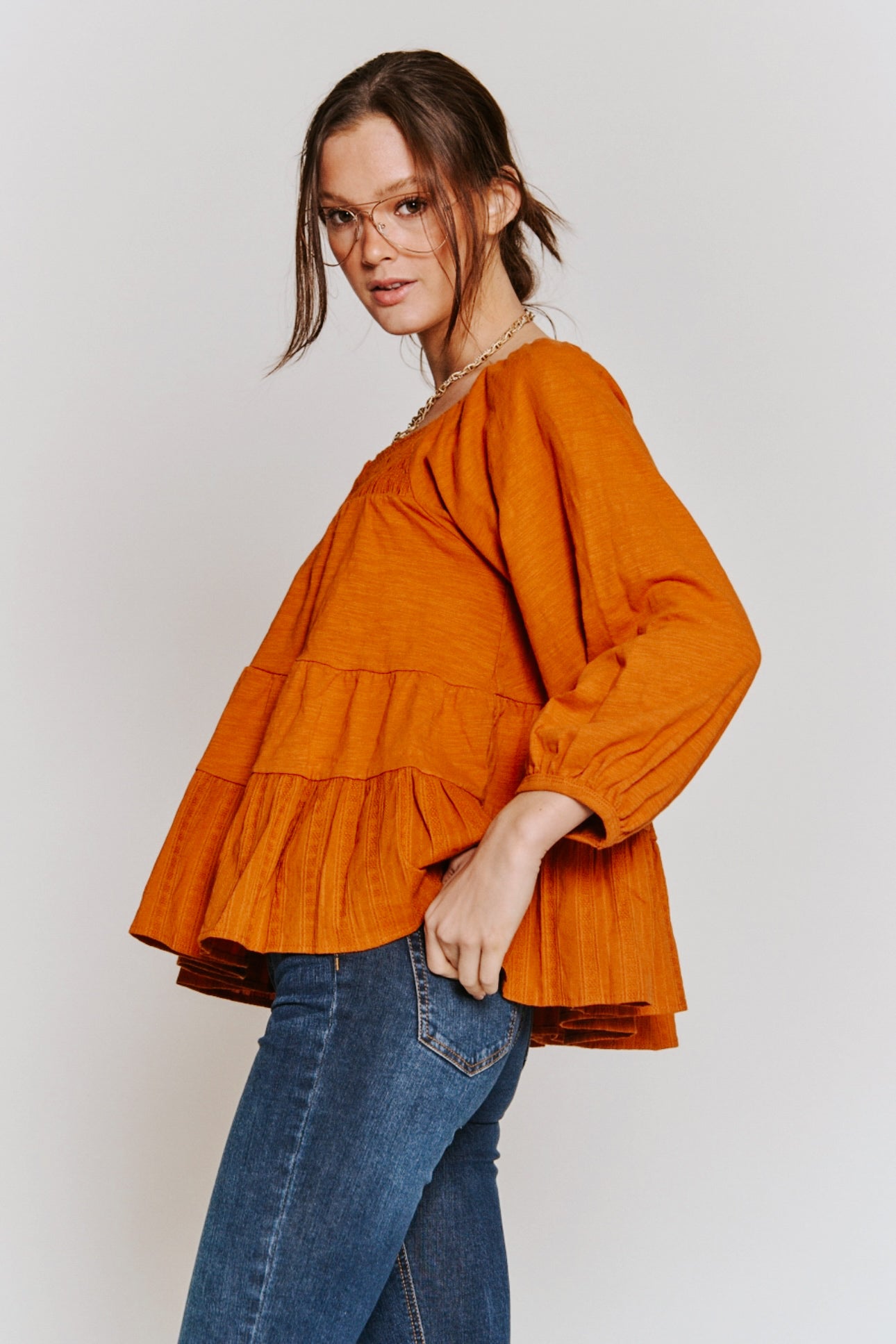 Woman wearing rust-colored ruched babydoll top with long sleeves paired with blue denim jeans against plain background