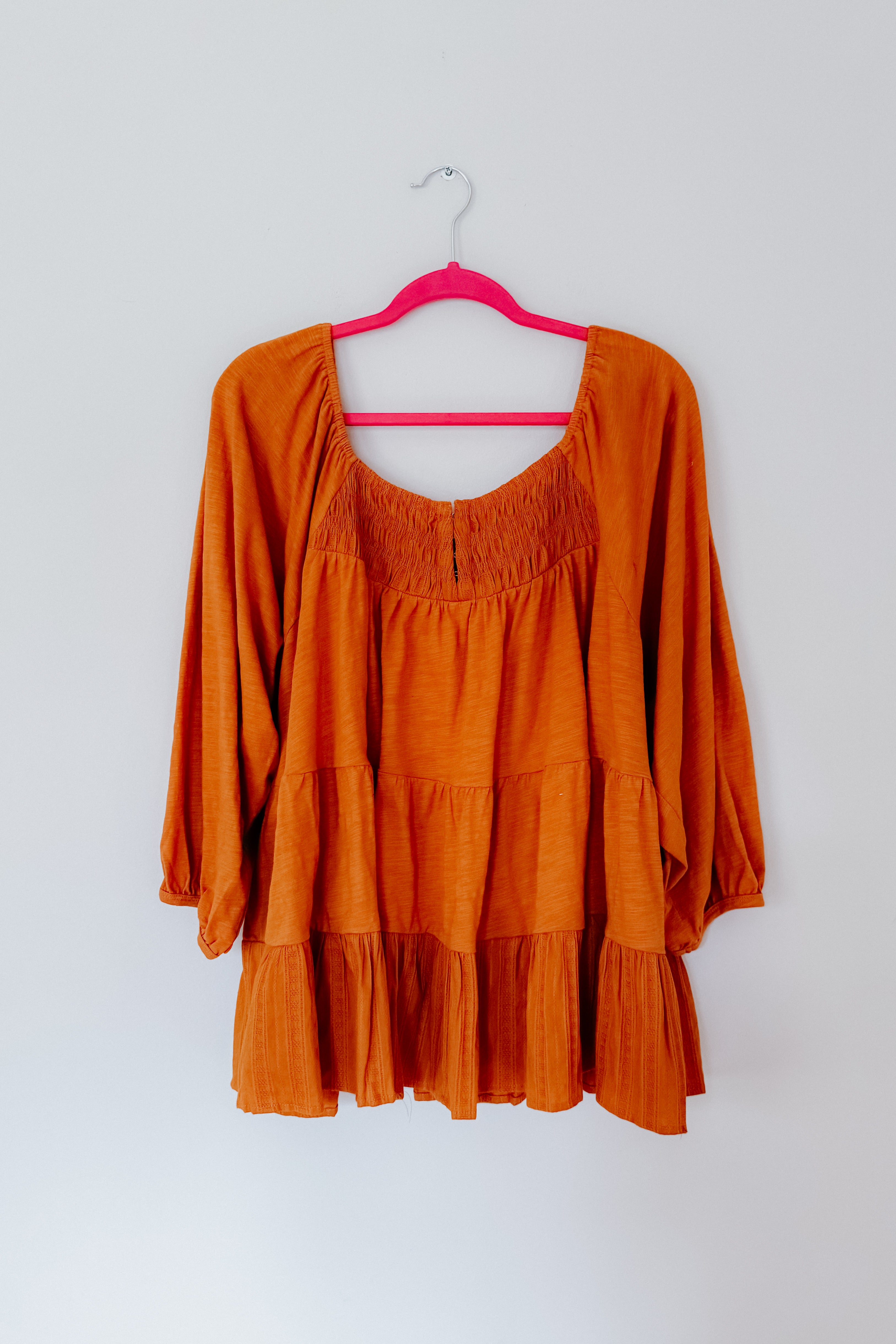 Rust-colored babydoll top with ruched neckline and long sleeves hanging on bright pink hanger against white wall