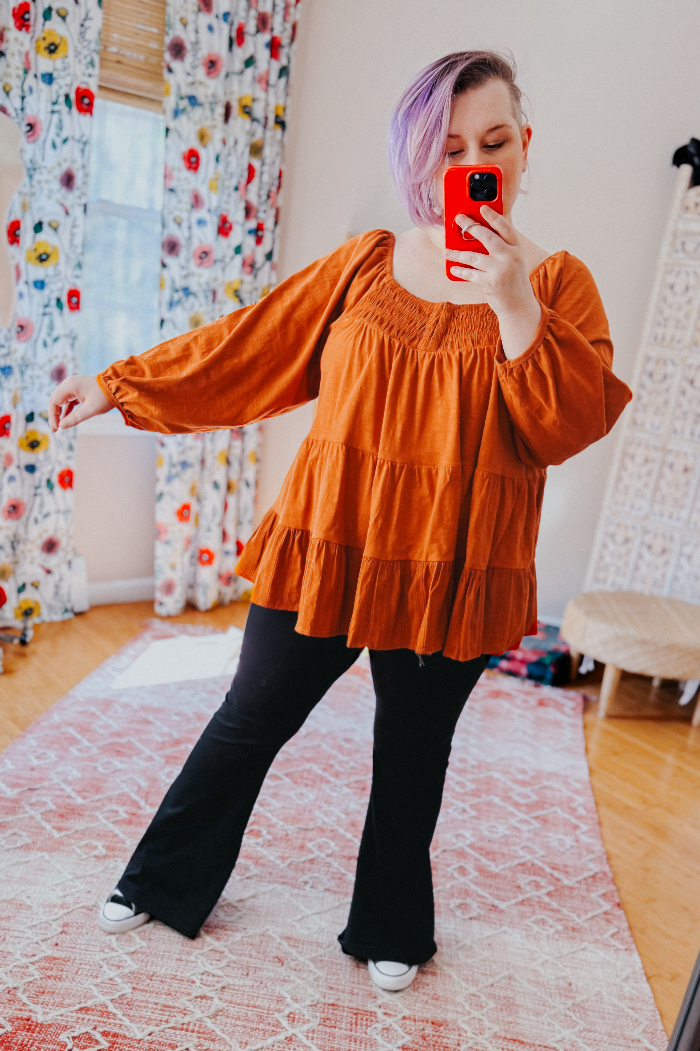 Woman with purple hair wearing rust-colored ruched babydoll top and black pants taking mirror selfie indoors