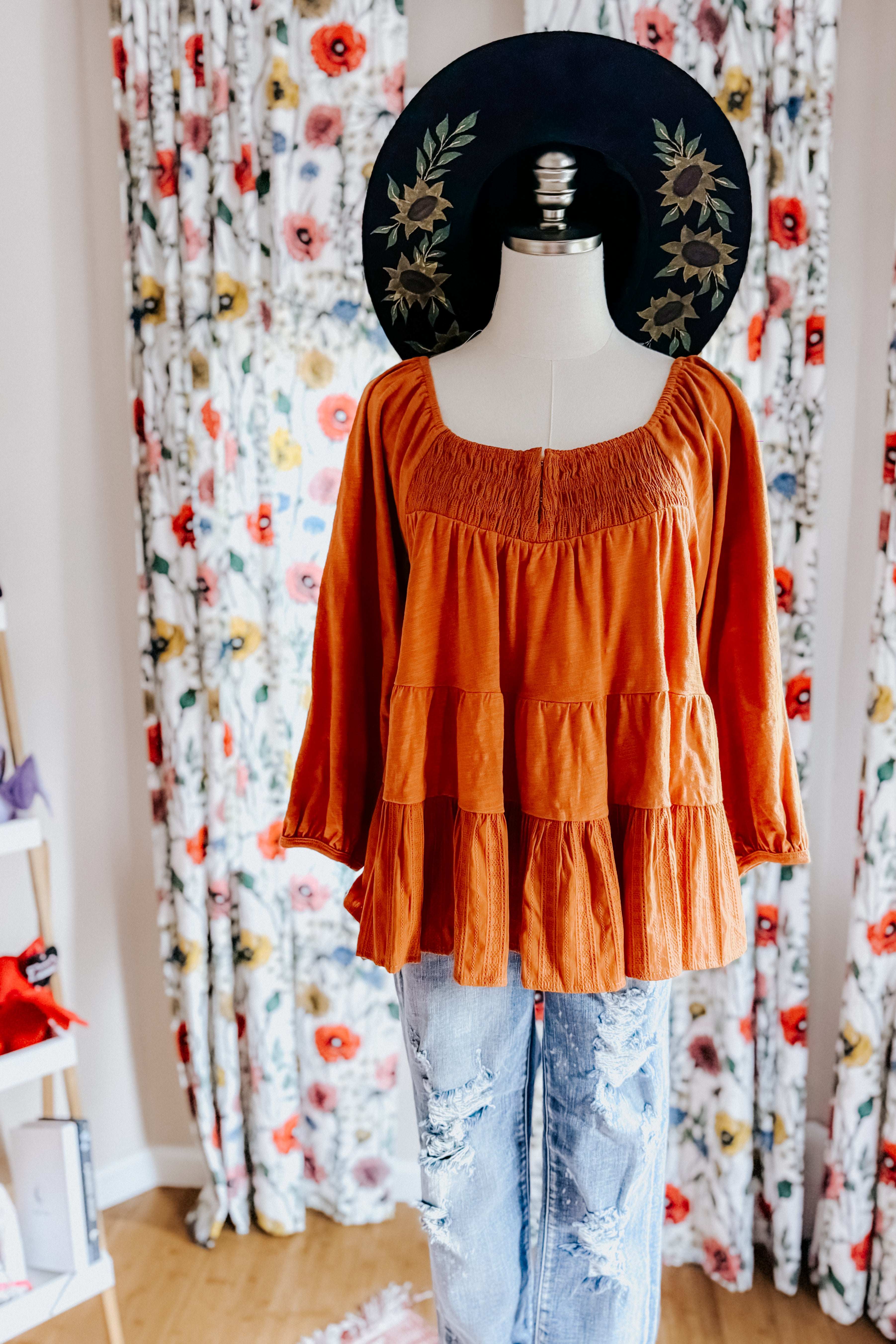 Rust-colored ruched babydoll top styled on mannequin with distressed jeans and black sunflower-embroidered hat