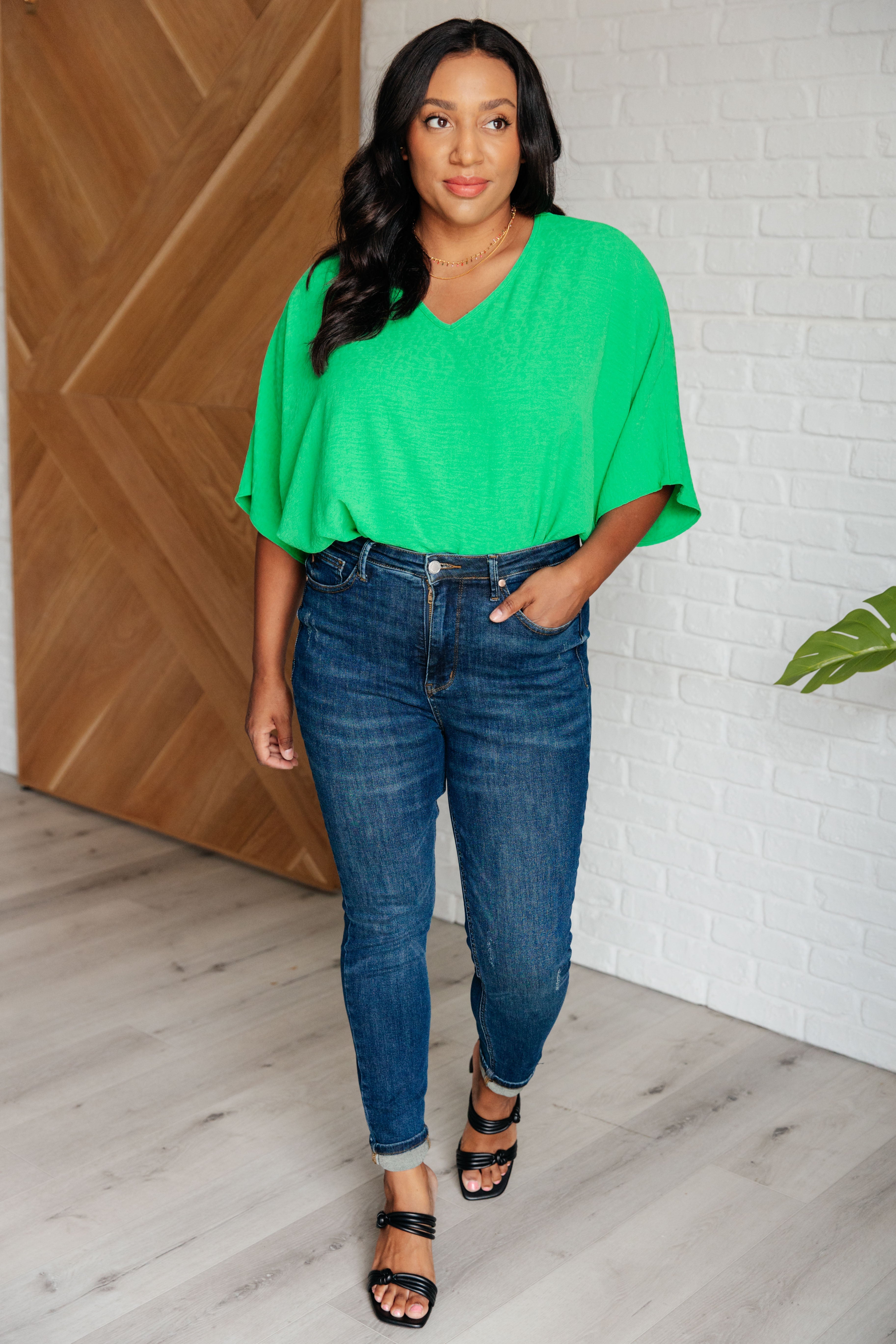 Woman wearing bright green relaxed-fit V-neck top paired with cuffed dark blue skinny jeans and black sandals indoors
