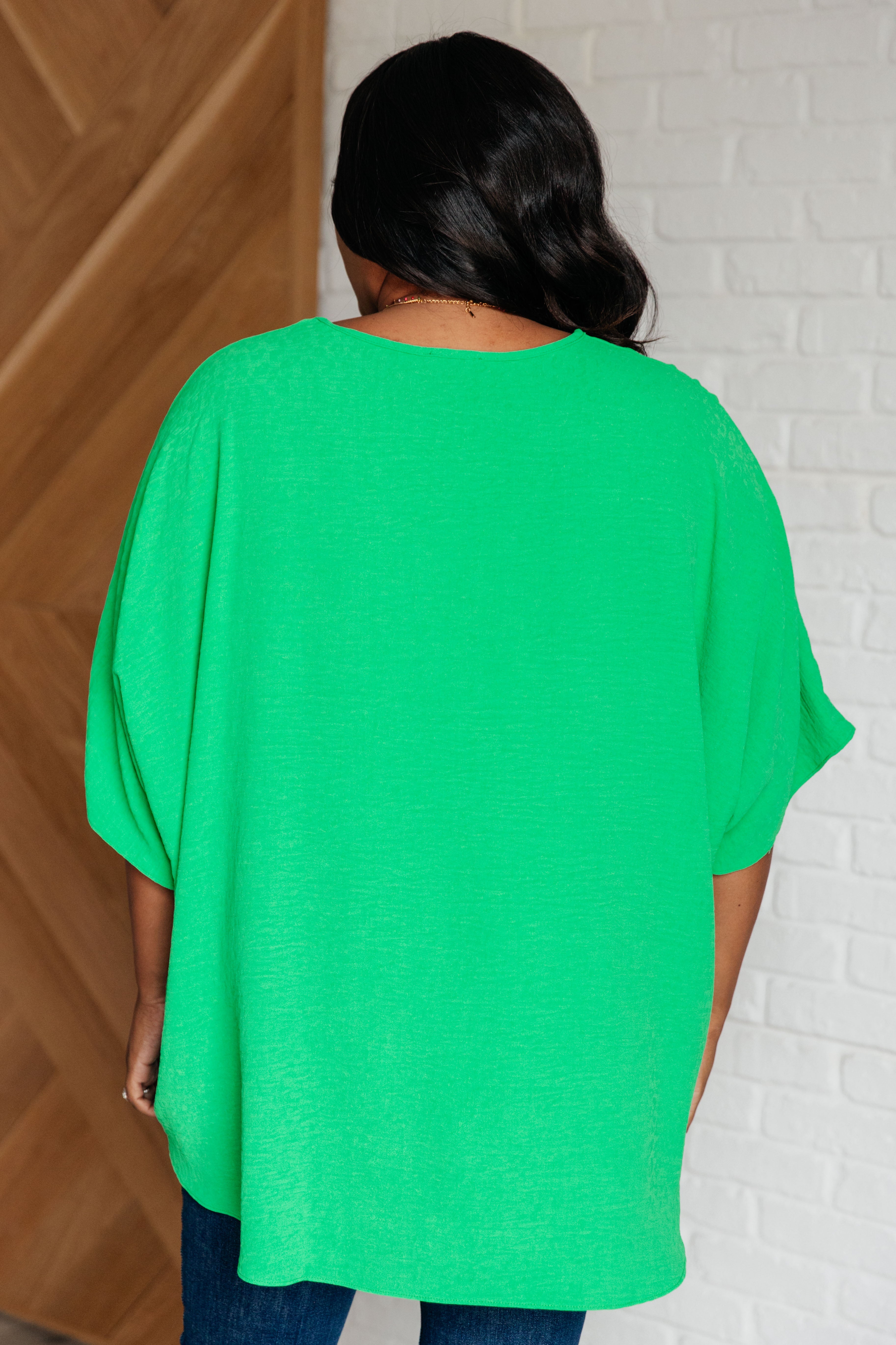 Plus-size woman wearing bright green loose-fitting V-neck top standing with her back to the camera indoors