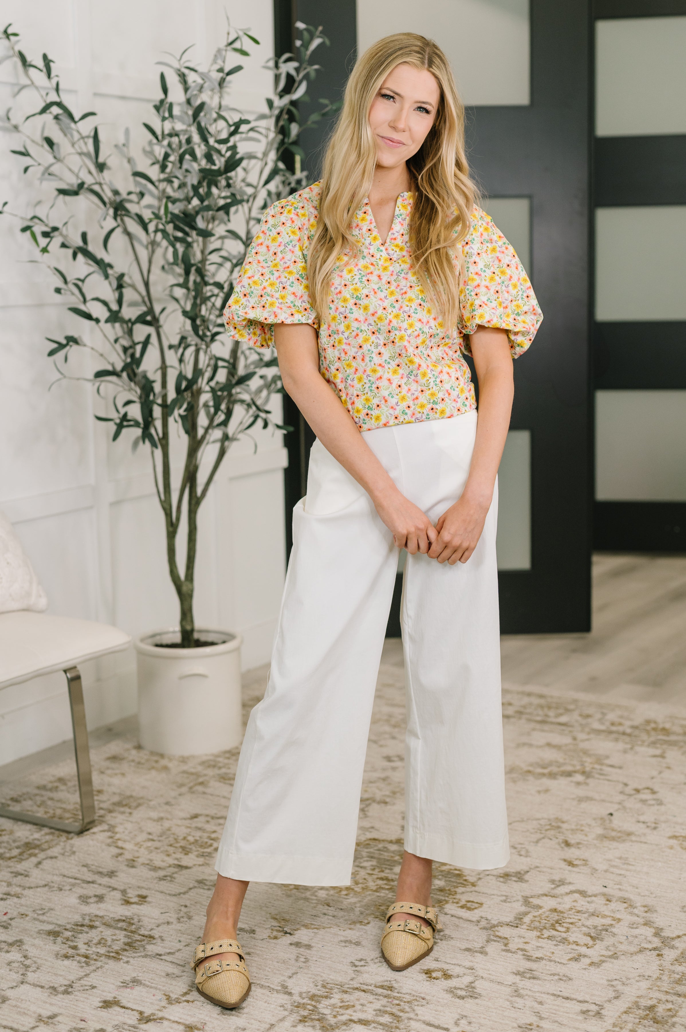 Woman wearing yellow floral balloon sleeve blouse paired with wide-leg white pants and studded flats indoors