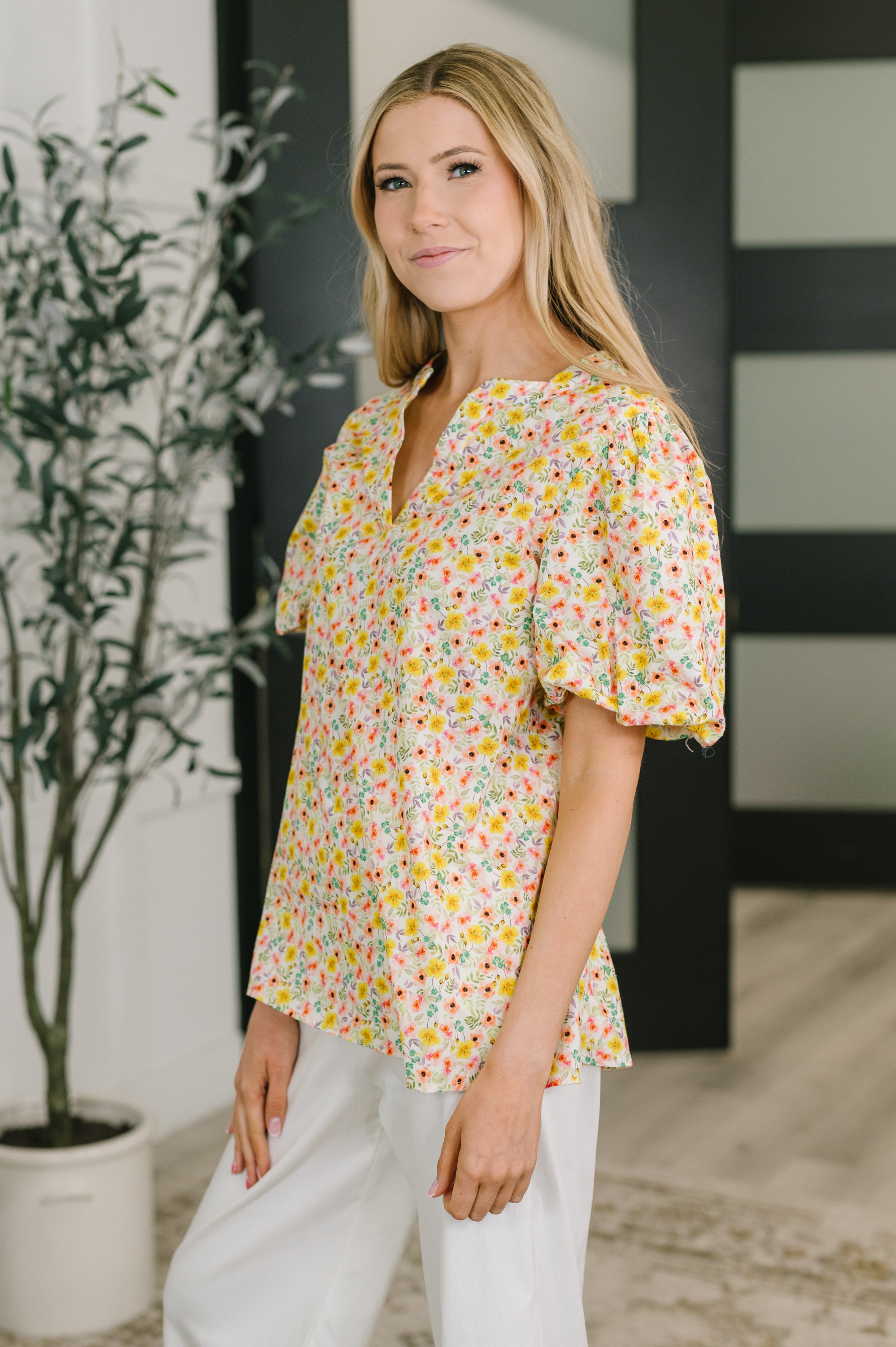 Woman wearing yellow floral balloon sleeve blouse with banded split neckline and white pants indoors