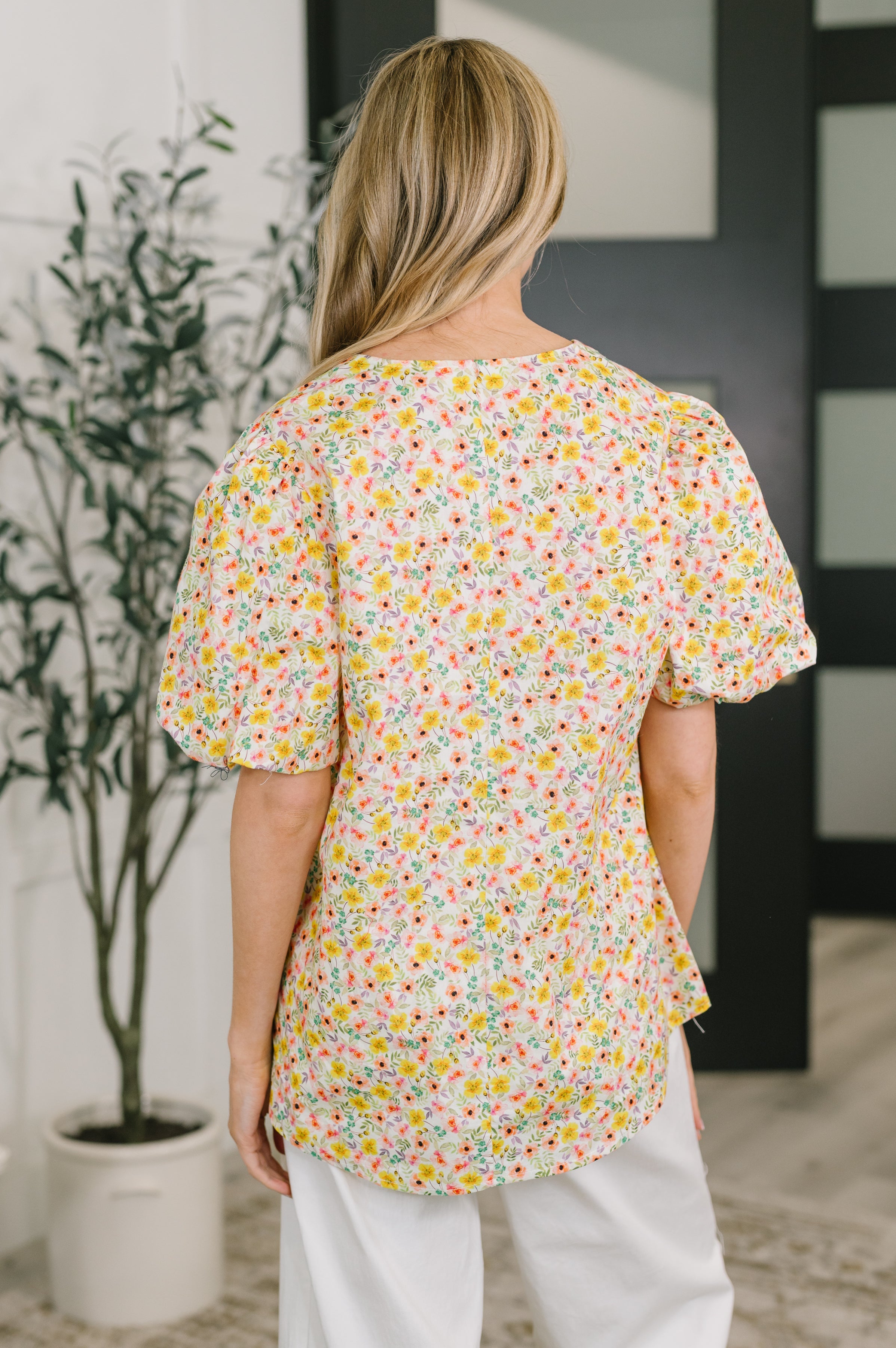 Back view of woman wearing yellow floral blouse with voluminous sleeves and relaxed fit paired with white pants indoors