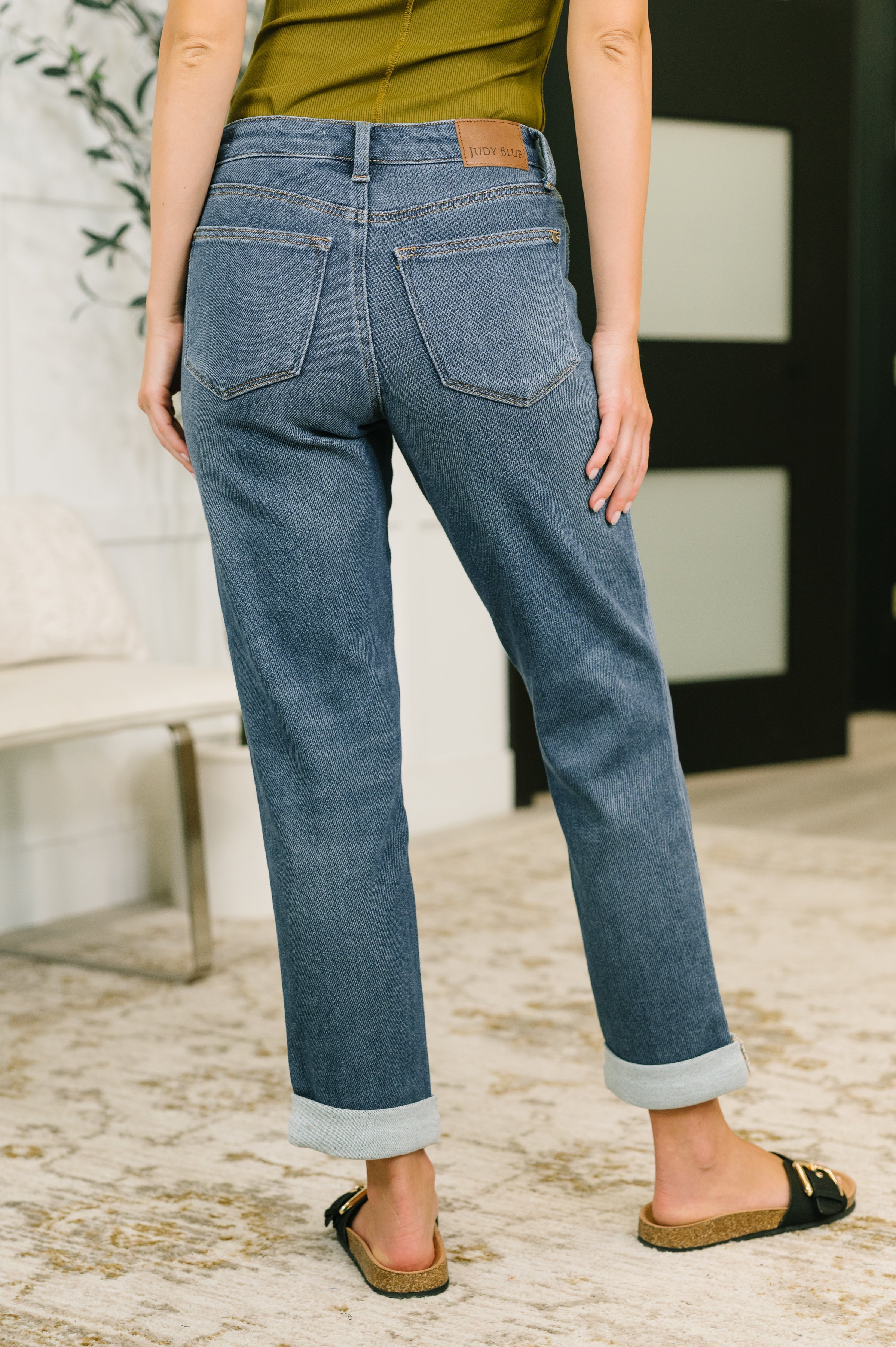 Back view of relaxed mid rise blue denim jeans with rolled cuffs worn with olive top and black slip sandals indoors